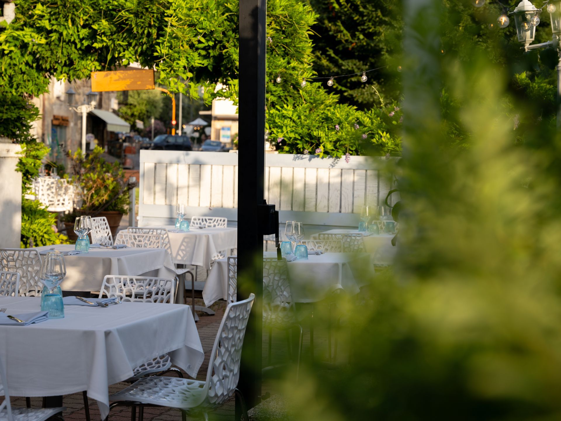 Tavoli da ristorante all'aperto con tovaglie bianche e bicchieri blu, alberi sullo sfondo.