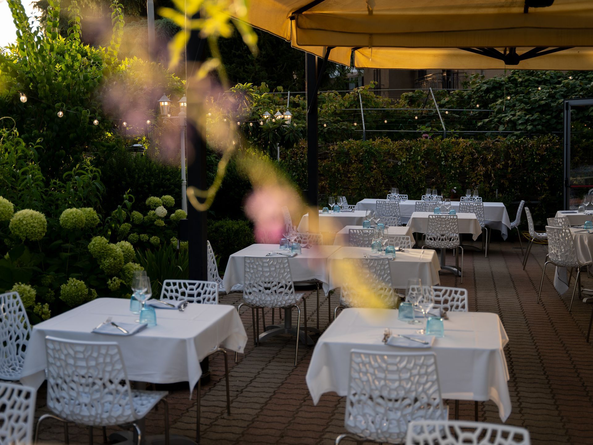 Ristorante all'aperto con tovaglie bianche, sedie e bicchieri sui tavoli sotto una tenda gialla, circondato dal verde.