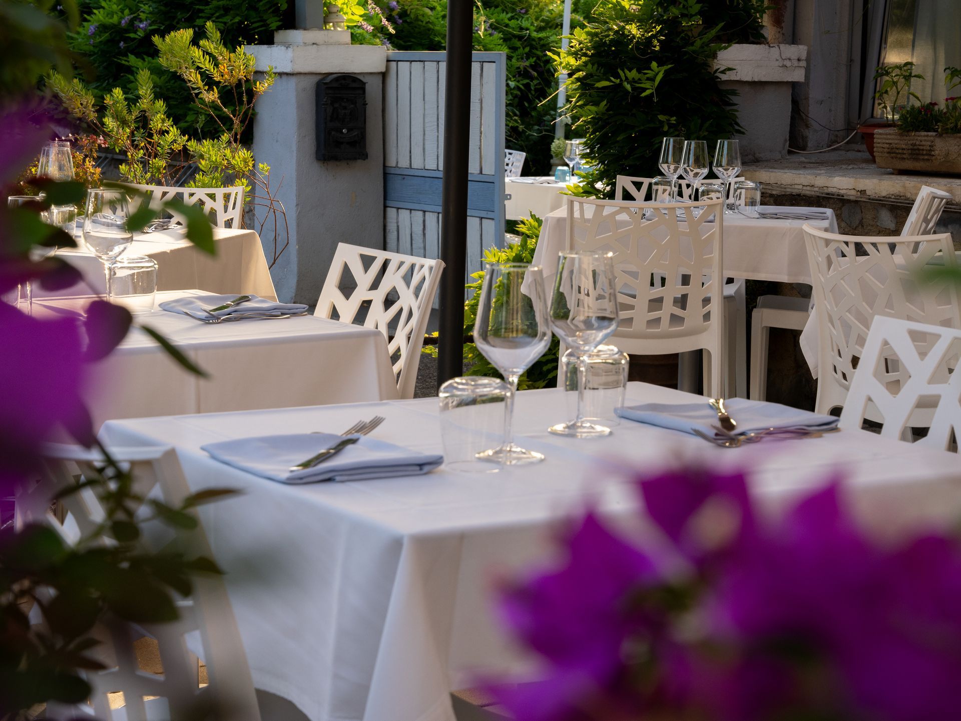 Tavoli da ristorante all'aperto apparecchiati con tovaglie bianche, sedie e bicchieri, circondati da piante e fiori viola.