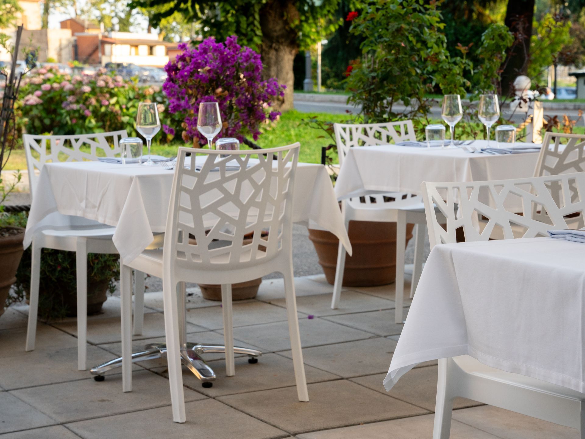 Tavoli da ristorante all'aperto con tovaglie bianche, sedie e bicchieri da vino, circondati da piante.
