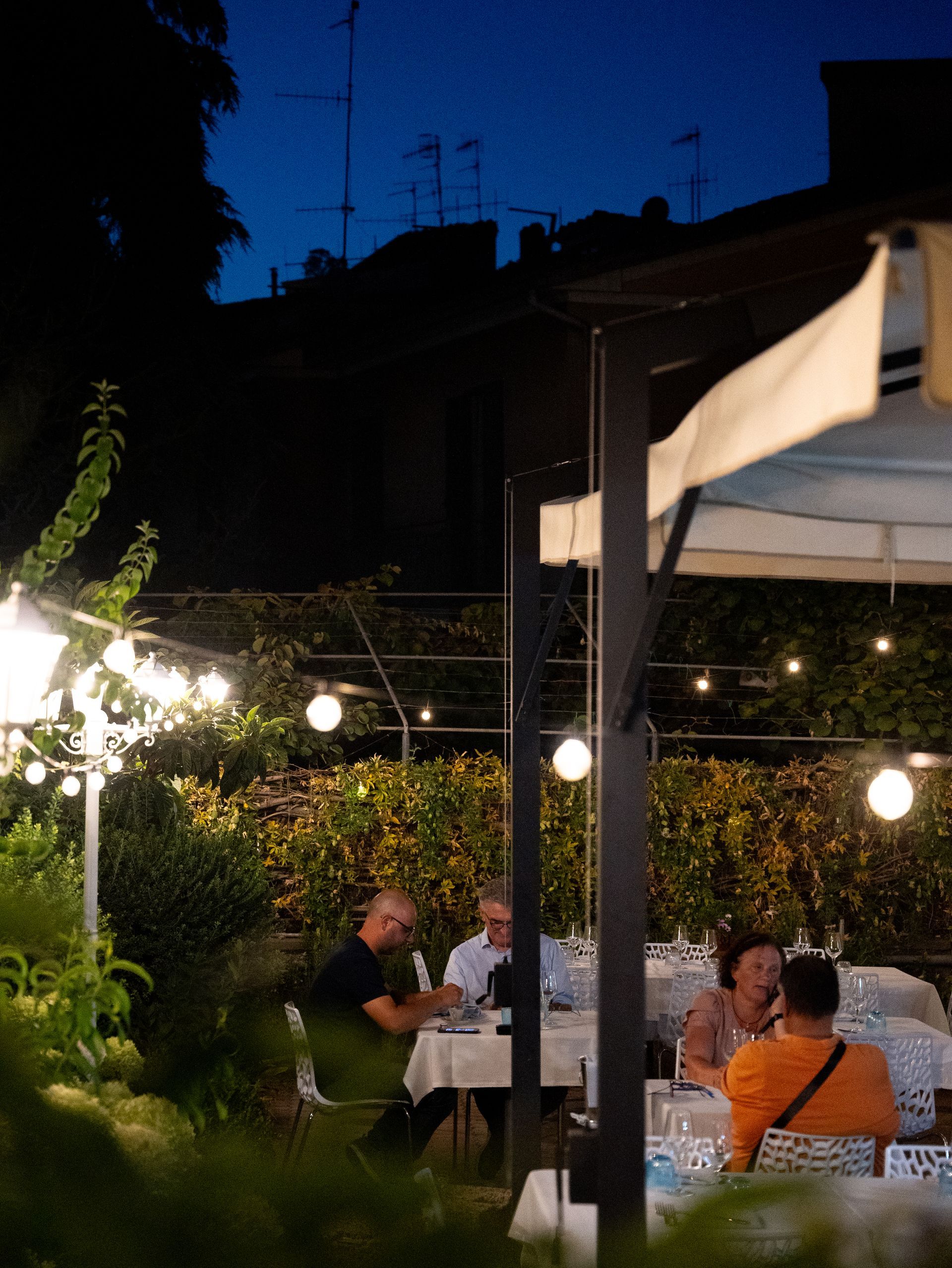 Ristorante all'aperto di notte, tavoli apparecchiati per i commensali sotto lucine, cielo blu scuro.