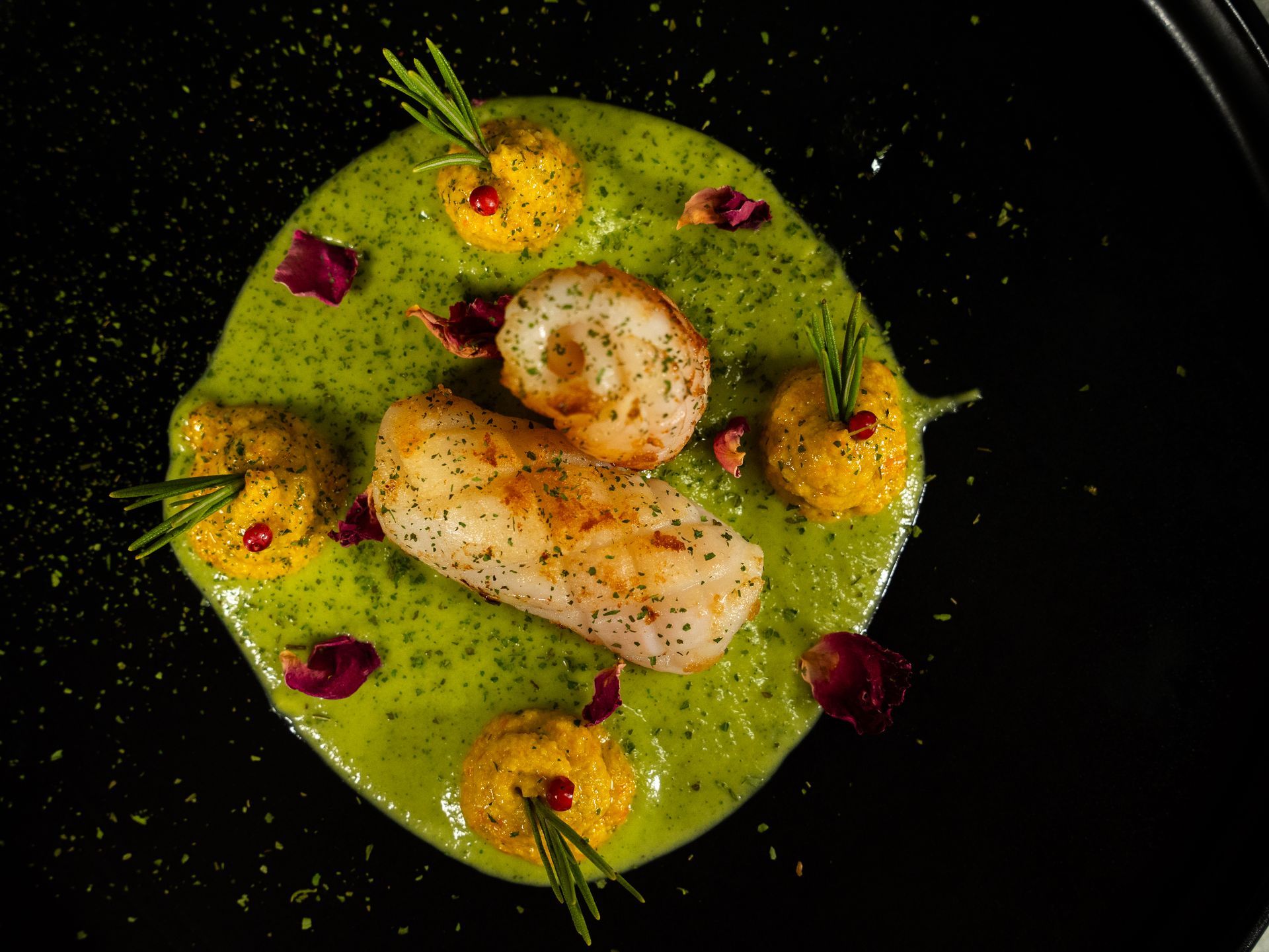Pesce scottato in padella con salsa verde, con polpette di patate gialle e guarnizione di rosmarino, su un piatto nero.