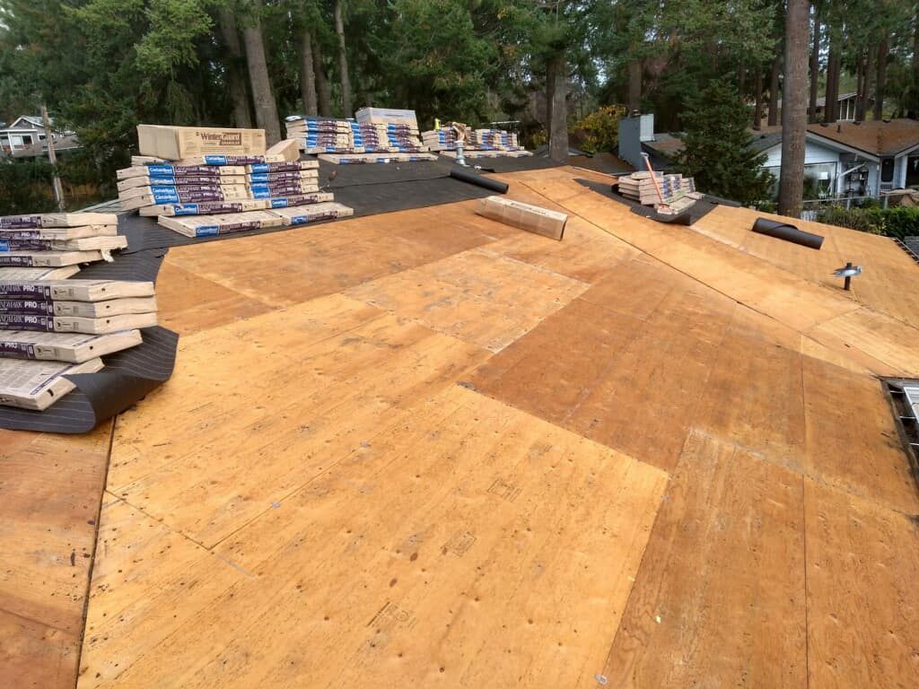 Roof deck under construction with plywood sheathing and stacked shingles beside trees and houses