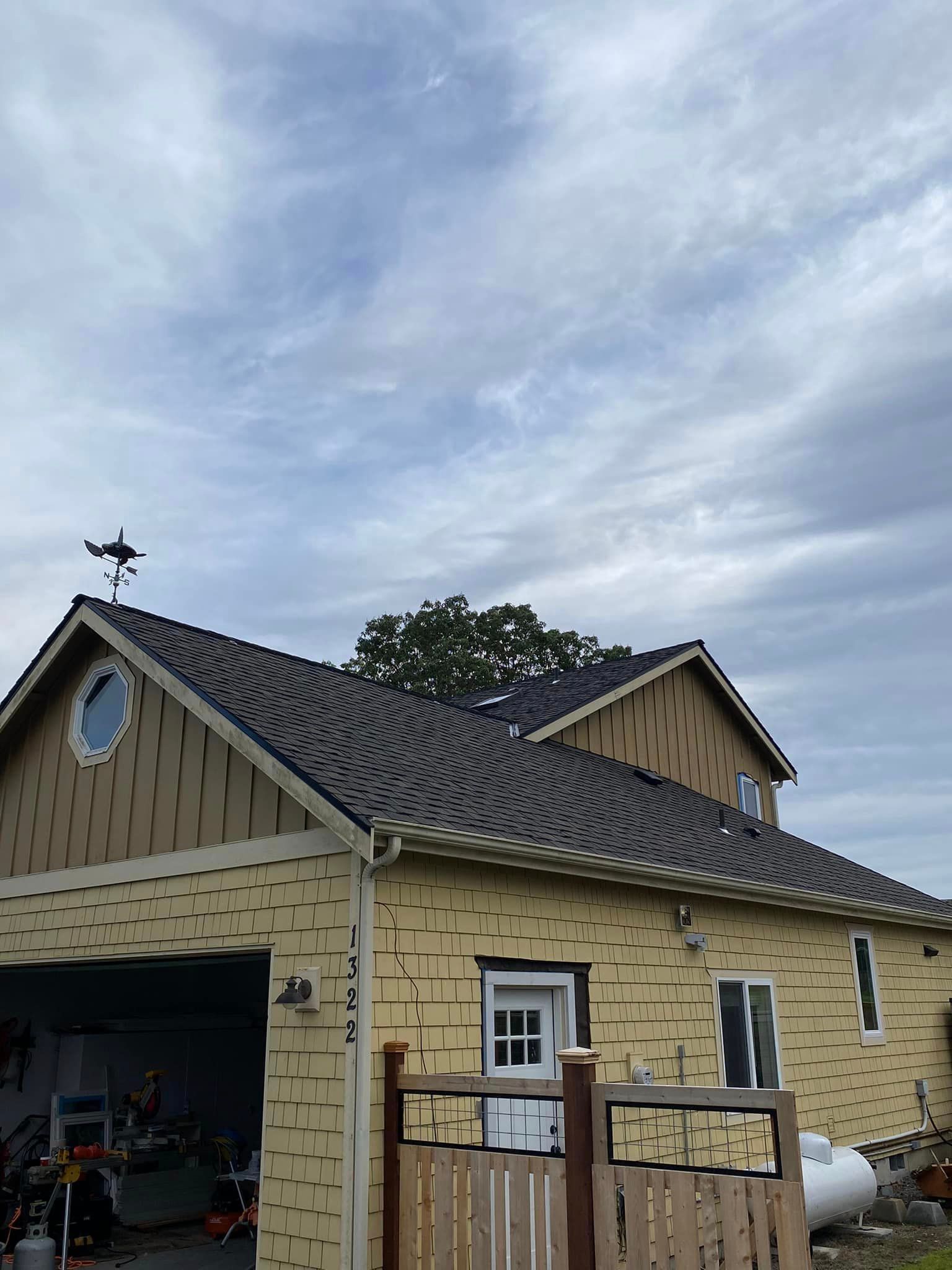 Stone garage with open bay, tan siding, gray shingled roof, and weather vane under a cloudy sky