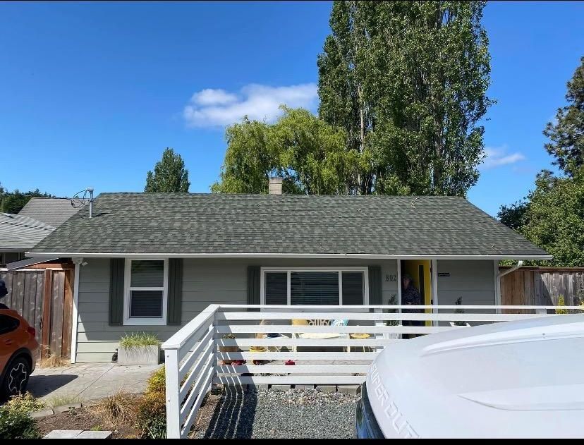 Small gray house with white fence and parked cars under a blue sky, with tall trees behind it