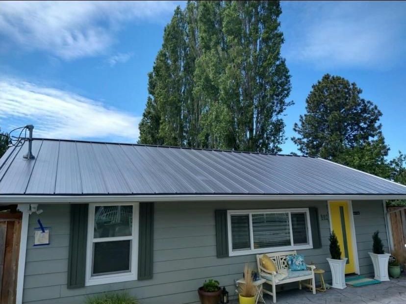Small gray house with a metal roof, porch, and tall trees against a blue sky.