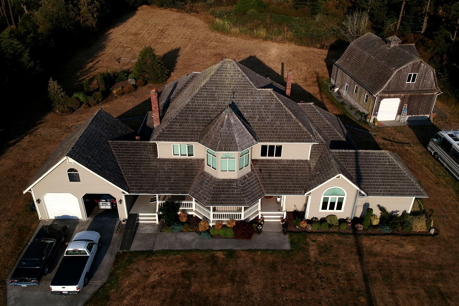 Aerial view of a large gray house with a driveway, garage, and parked vehicles in a rural setting