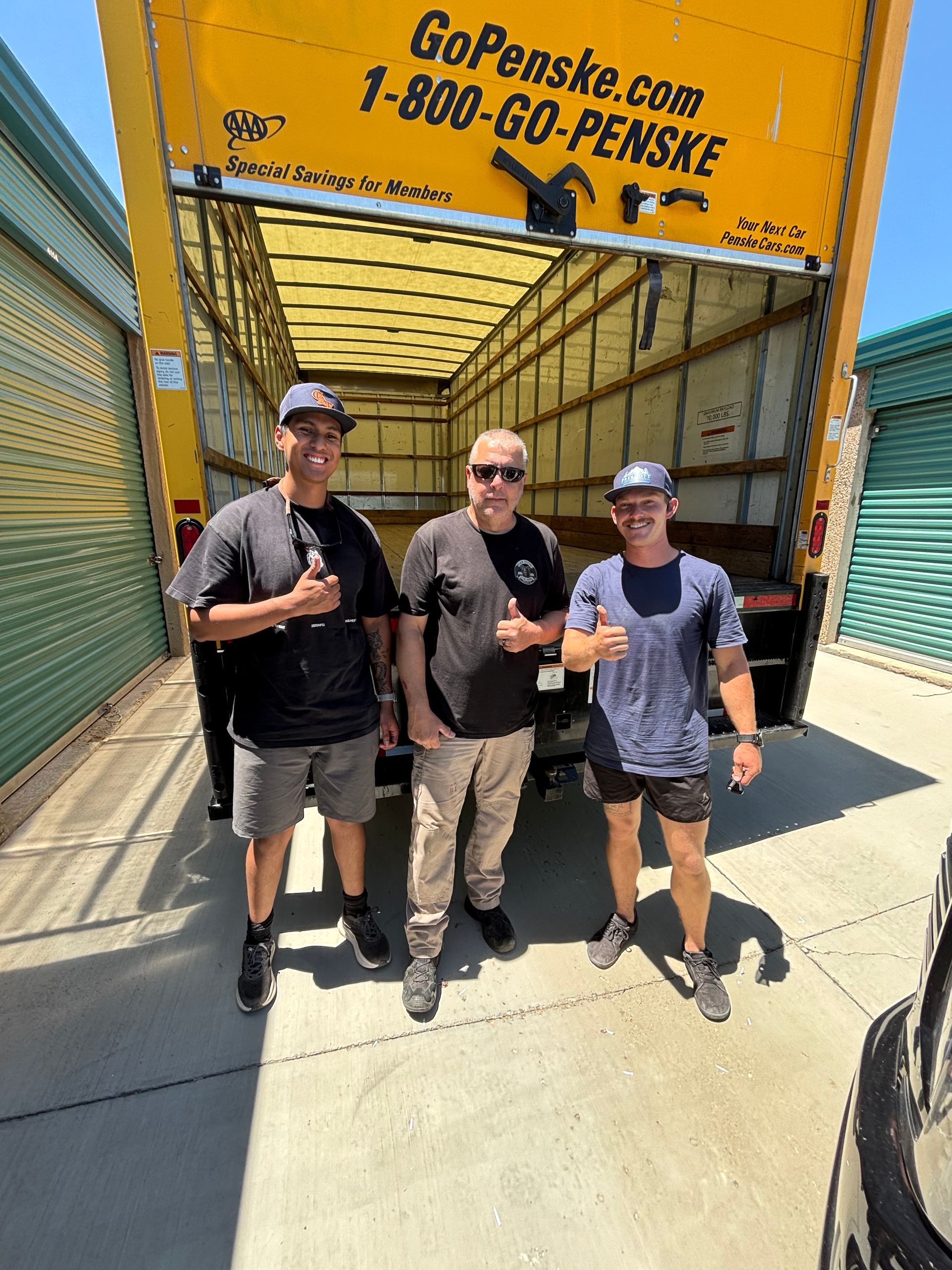 Three people posing with drinks under a yellow GoPenske.com truck trailer at a loading dock