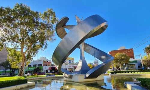 Gran escultura abstracta de metal en un parque soleado con árboles, césped y un estanque reflectante.