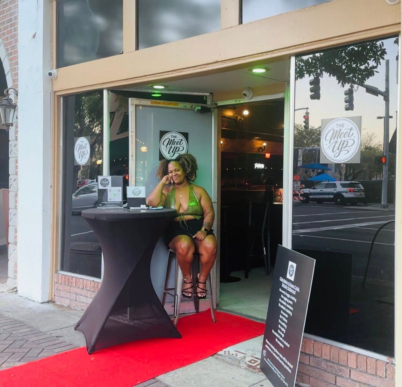 Woman seated at a small table outside a storefront on a red carpet, speaking into a microphone.