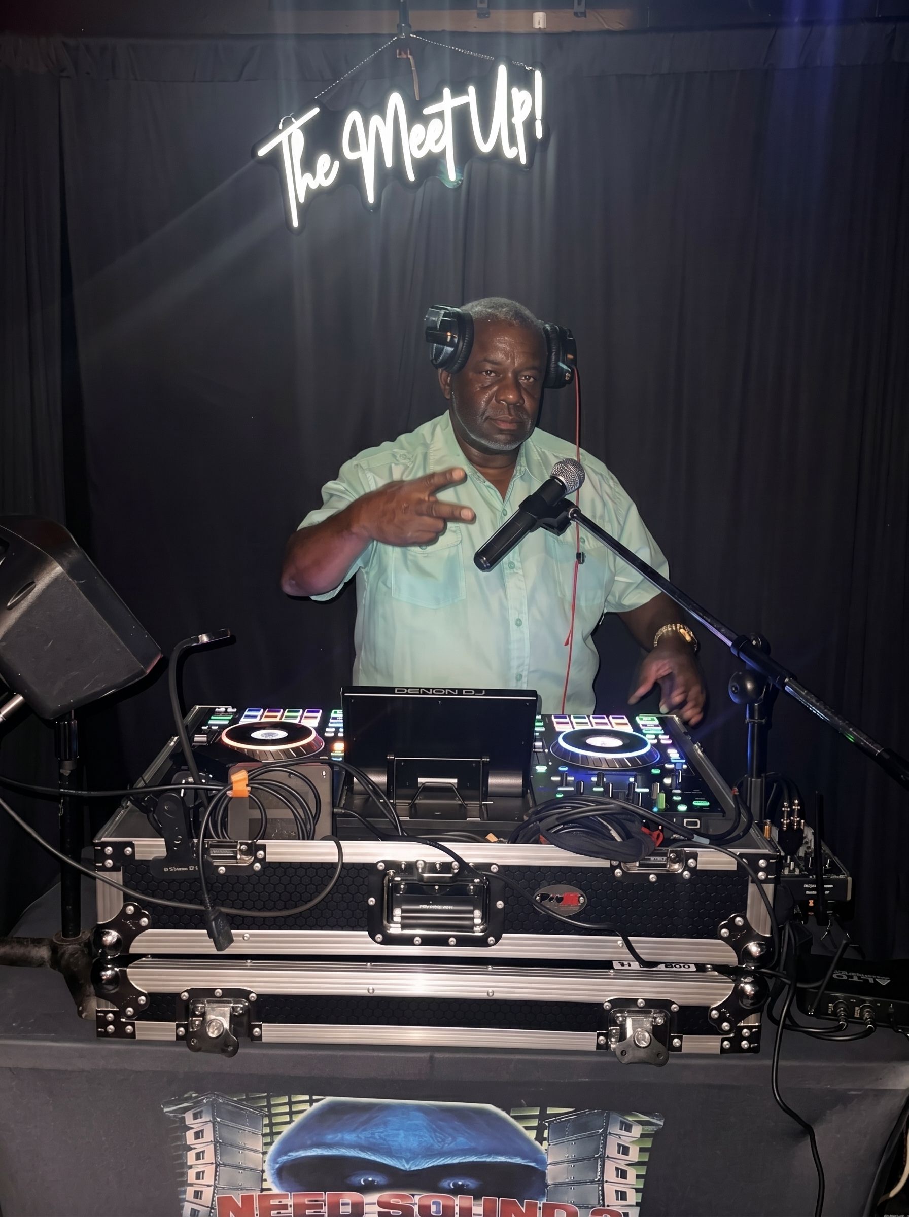 DJ performing behind a turntable setup on a dark stage, with one hand raised and a microphone nearby.