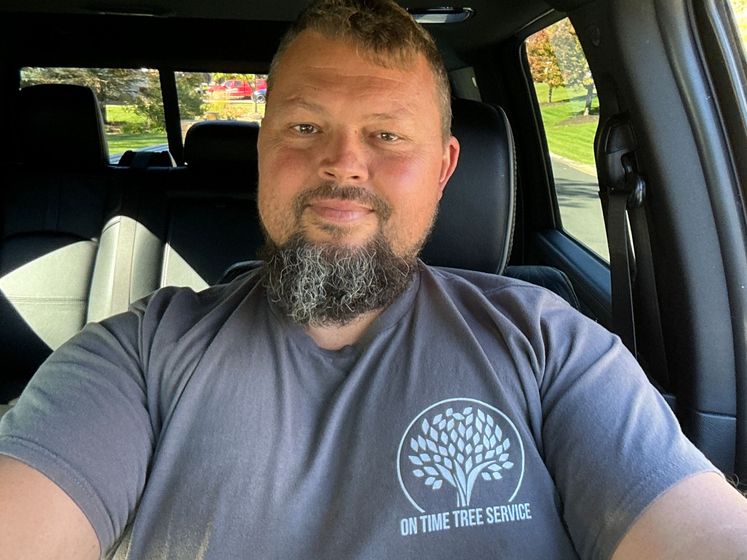 A man with a short beard sits in the driver's seat of a vehicle, wearing a gray t-shirt with a tree logo.