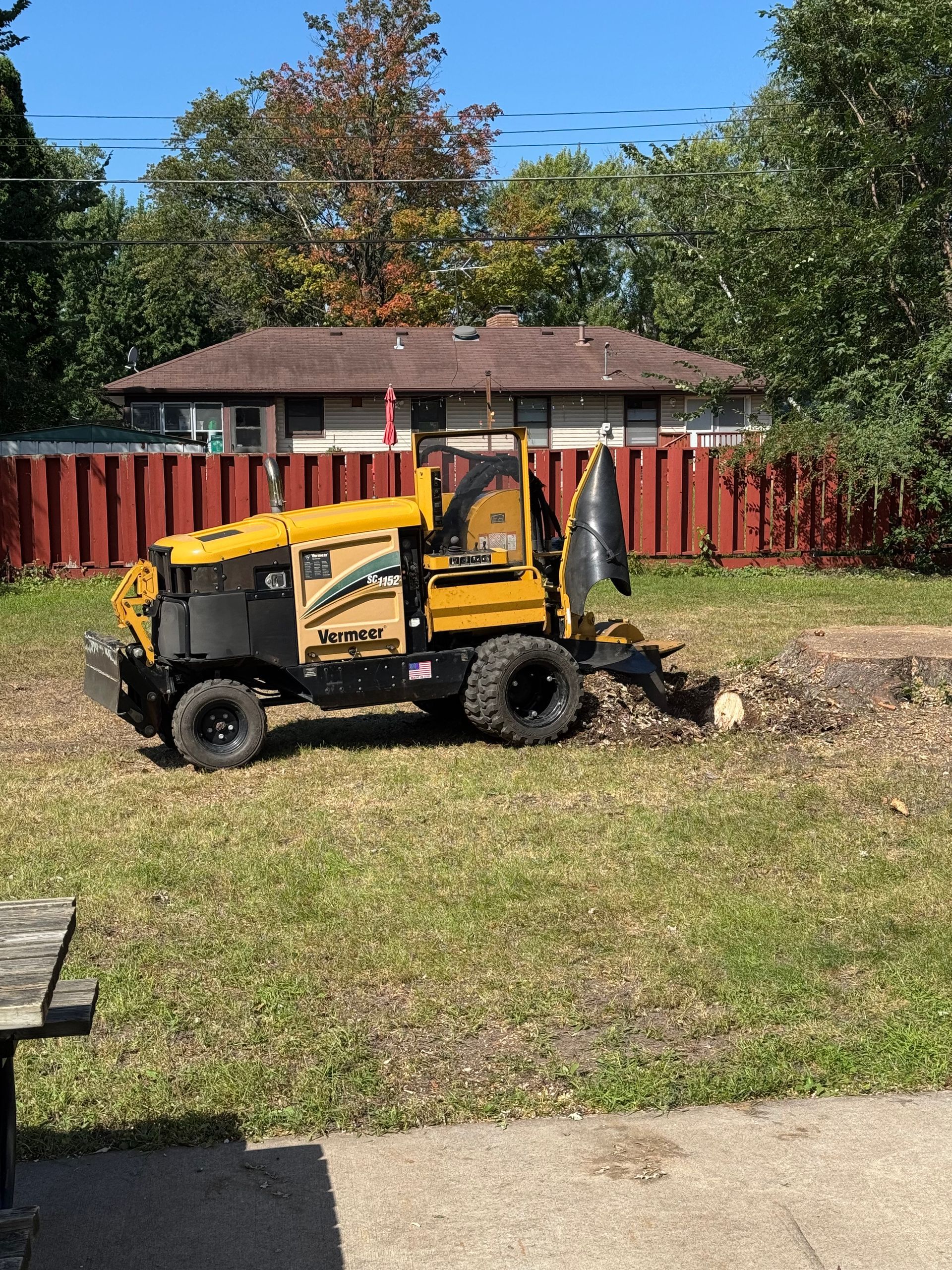 Yellow stump grinder on grass, grinding a tree stump in a backyard. House and trees in background.