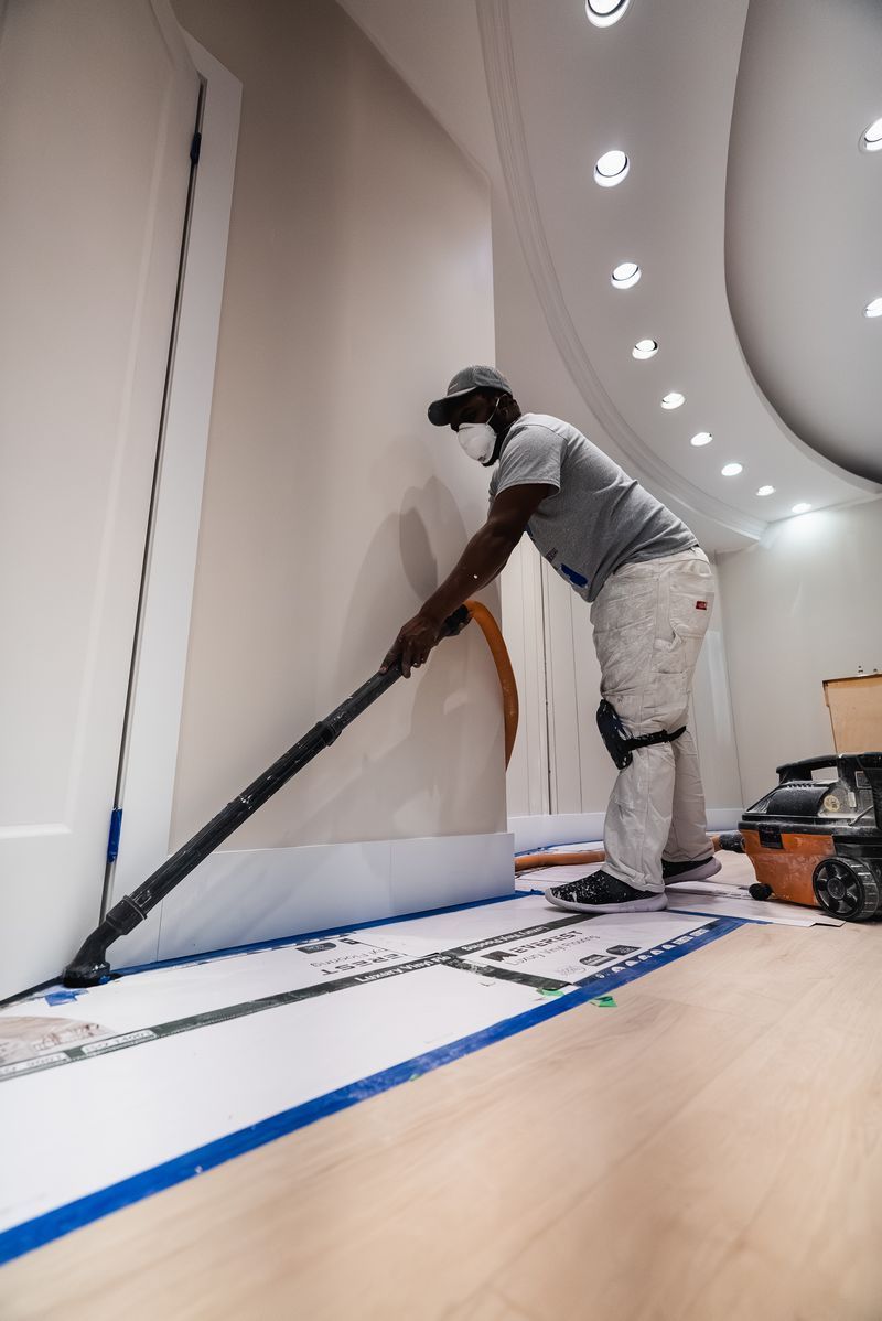 Person in protective gear sanding a wall. The room is light with curved ceiling and hardwood floor covered with paper.