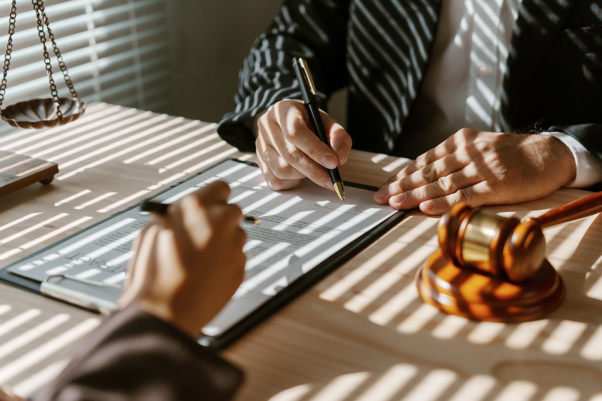 Hands signing legal documents at a desk with a gavel and striped sunlight shadows