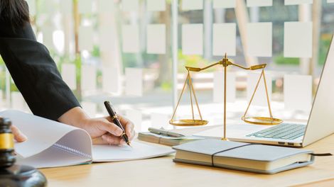 Lawyer signing papers beside a brass scales of justice and a laptop in a bright office Lawyer signing papers beside a brass scales of justice and a laptop in a bright office