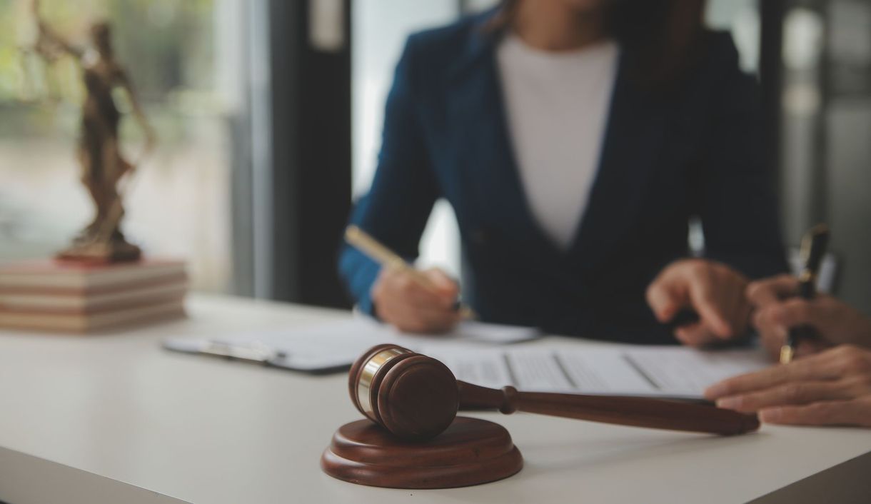 Judge’s gavel on a desk in front of a blurred person signing documents in an office Judge’s gavel on a desk in front of a blurred person signing documents in an office