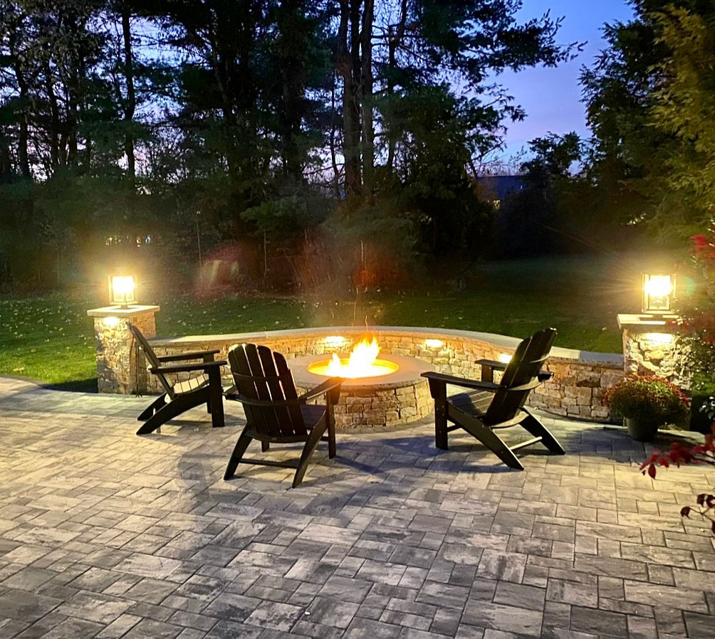 Backyard fire pit with four black chairs on a stone patio at dusk, lit by warm outdoor lights