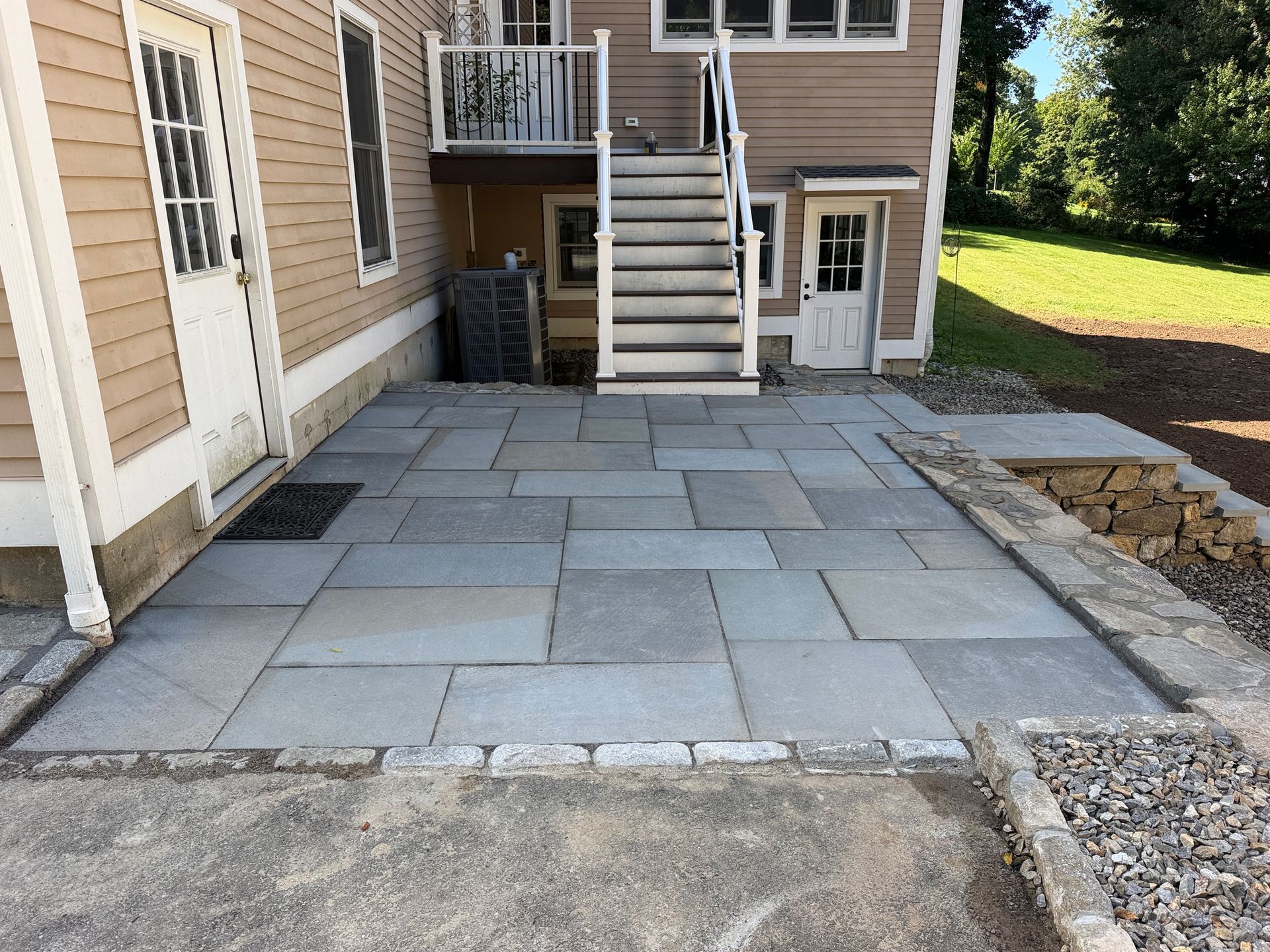 Stone patio with large gray pavers beside a house, leading to outdoor stairs and a basement door.