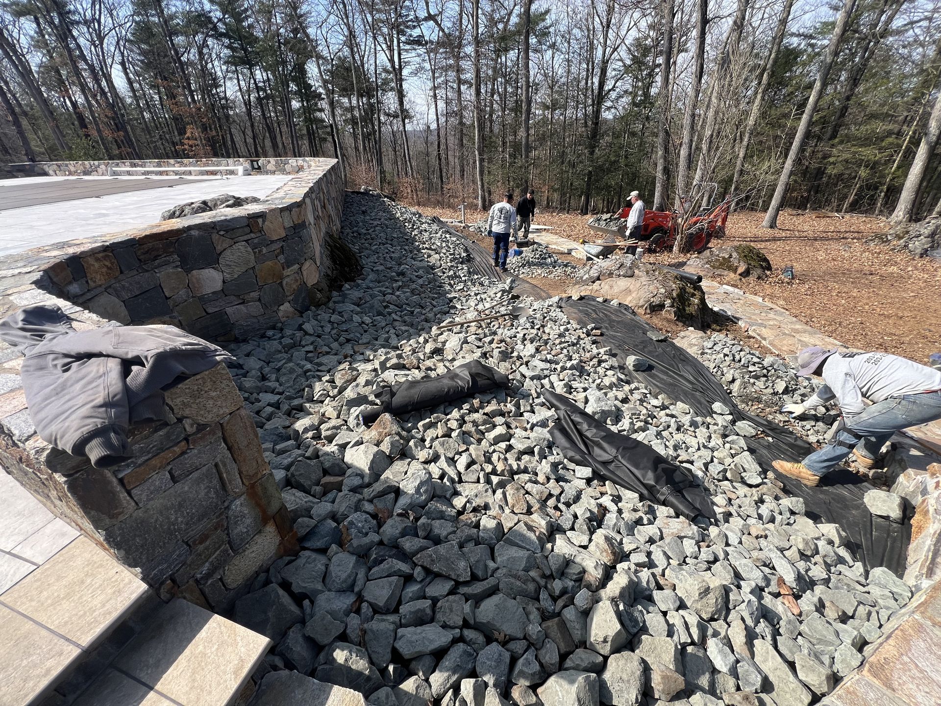 Outdoor stone construction site with workers around a curved retaining wall and gravel-filled area