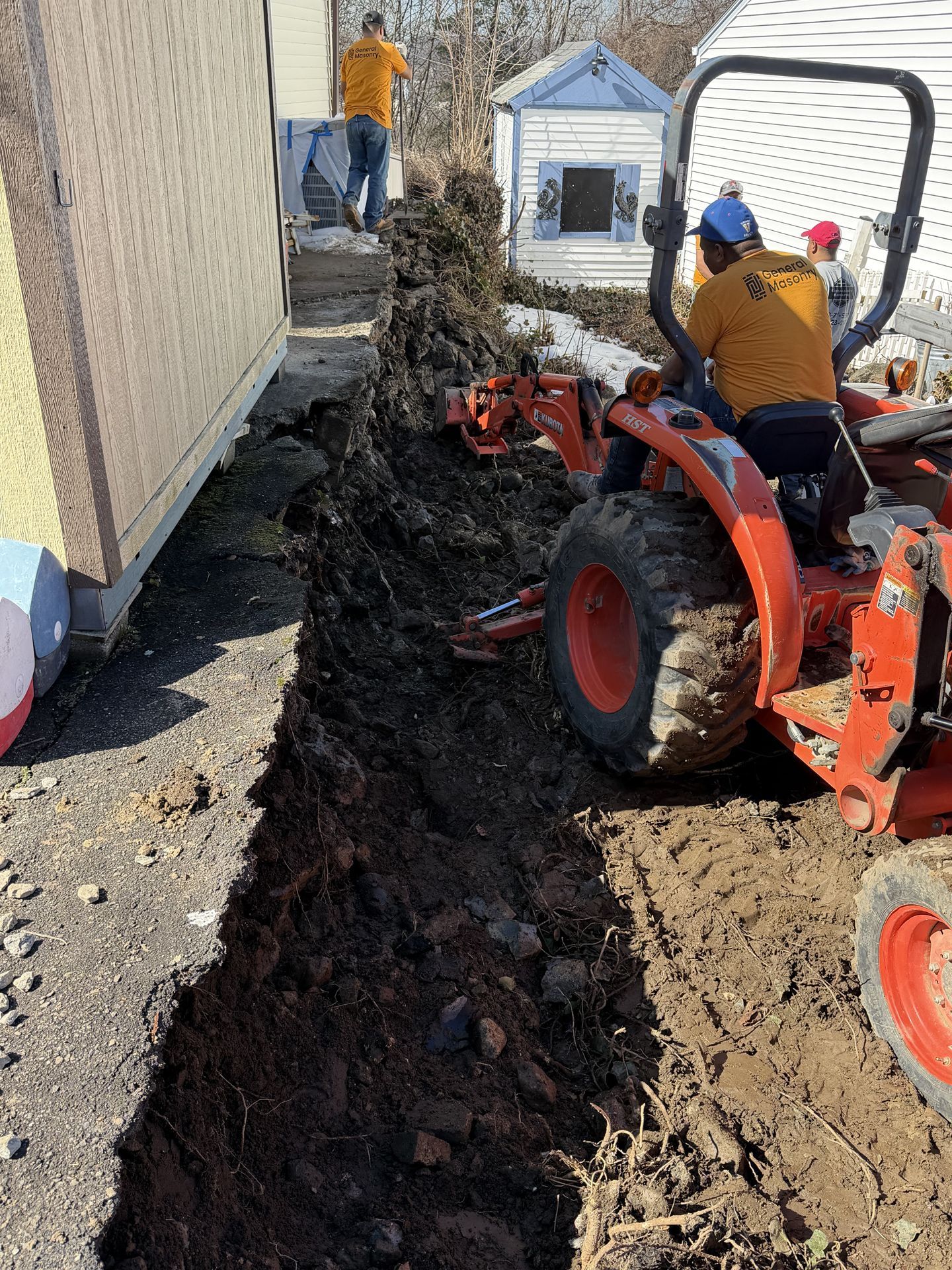 Construction crew using a tractor to dig a trench beside a house, with mud piled along the path.