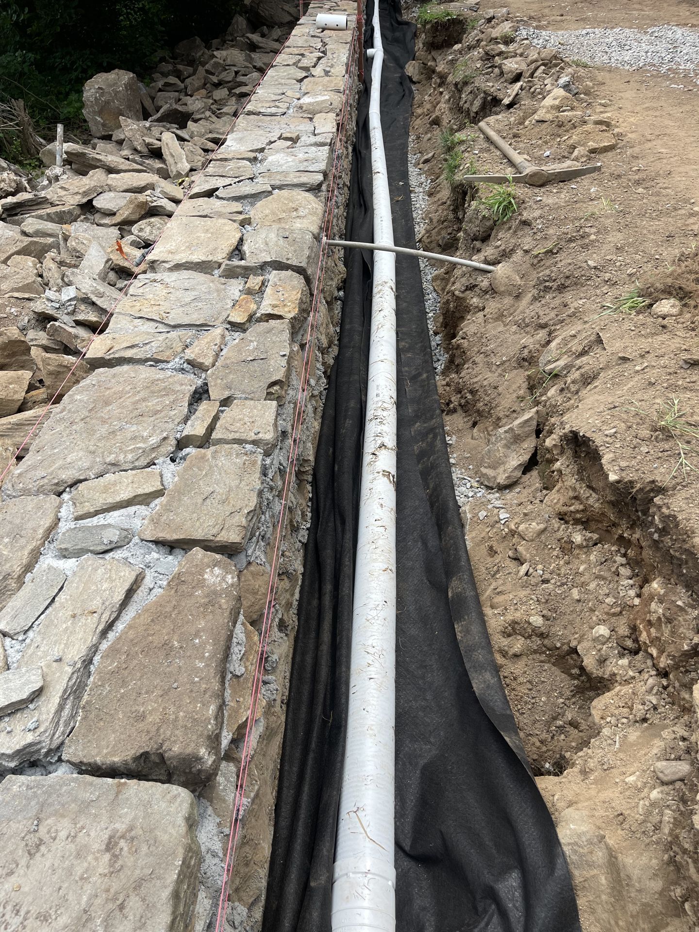 Stone retaining wall beside a narrow trench with a white pipe lined in black fabric on a dirt slope