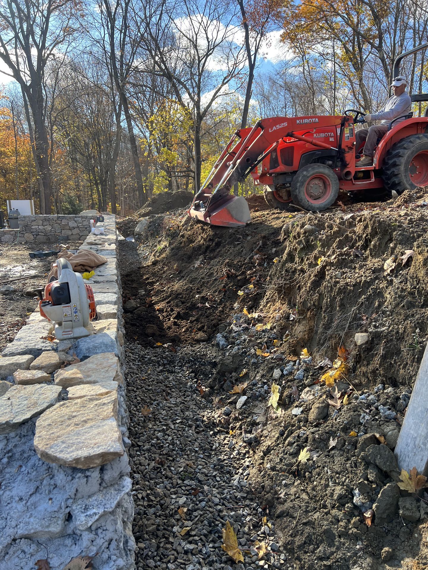Red tractor digging a trench beside a stone wall in a wooded outdoor area