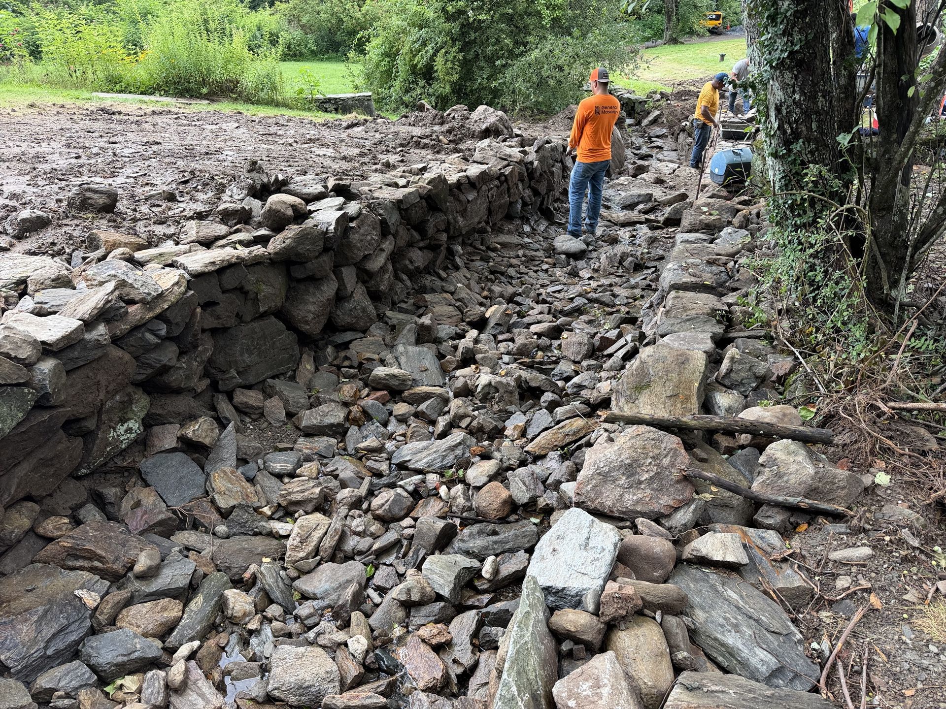 Collapsed rocky path with two workers inspecting flood damage near a wooded stream