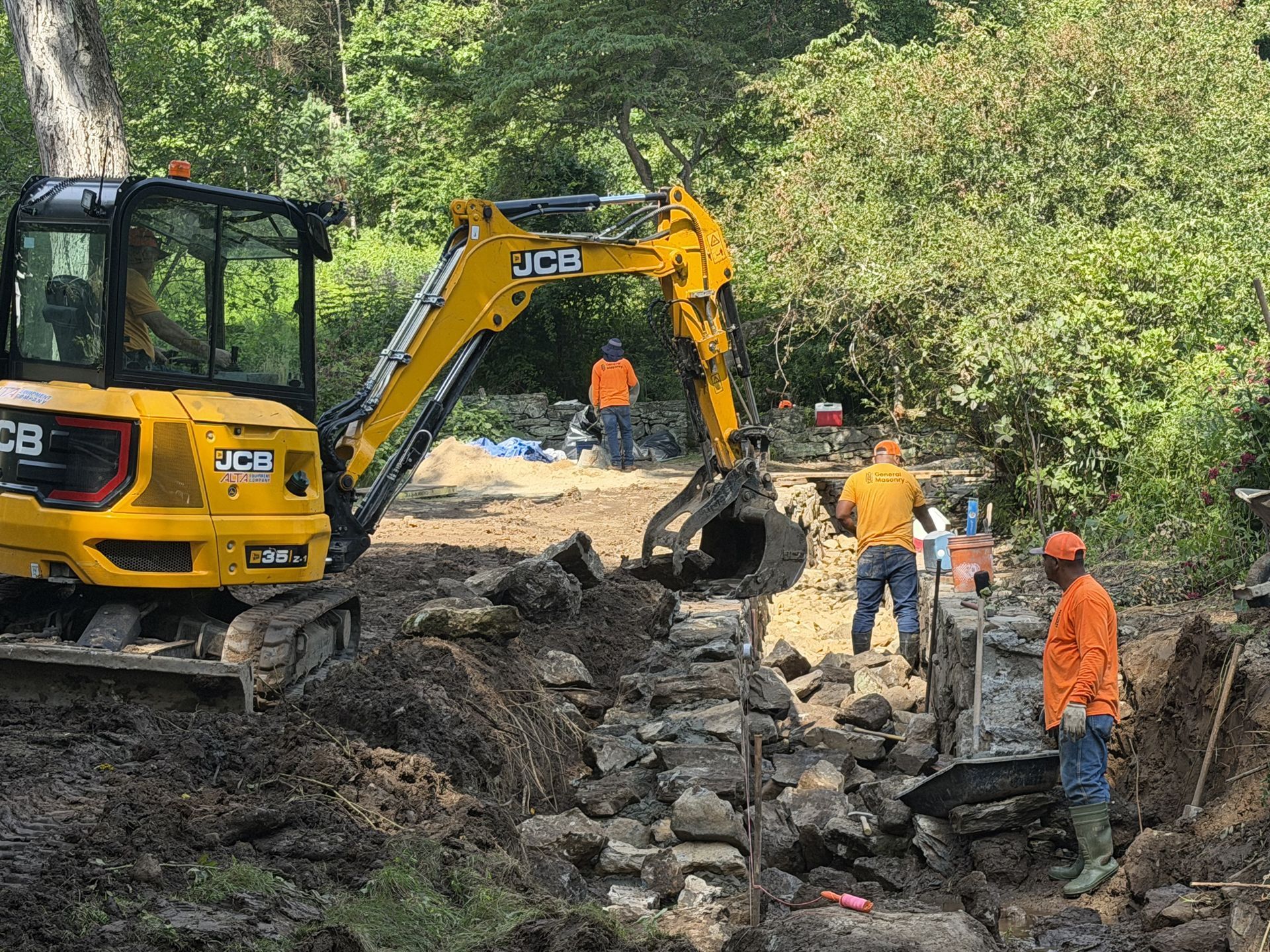 Yellow excavator and workers clearing rubble from a muddy roadside ditch in a wooded area