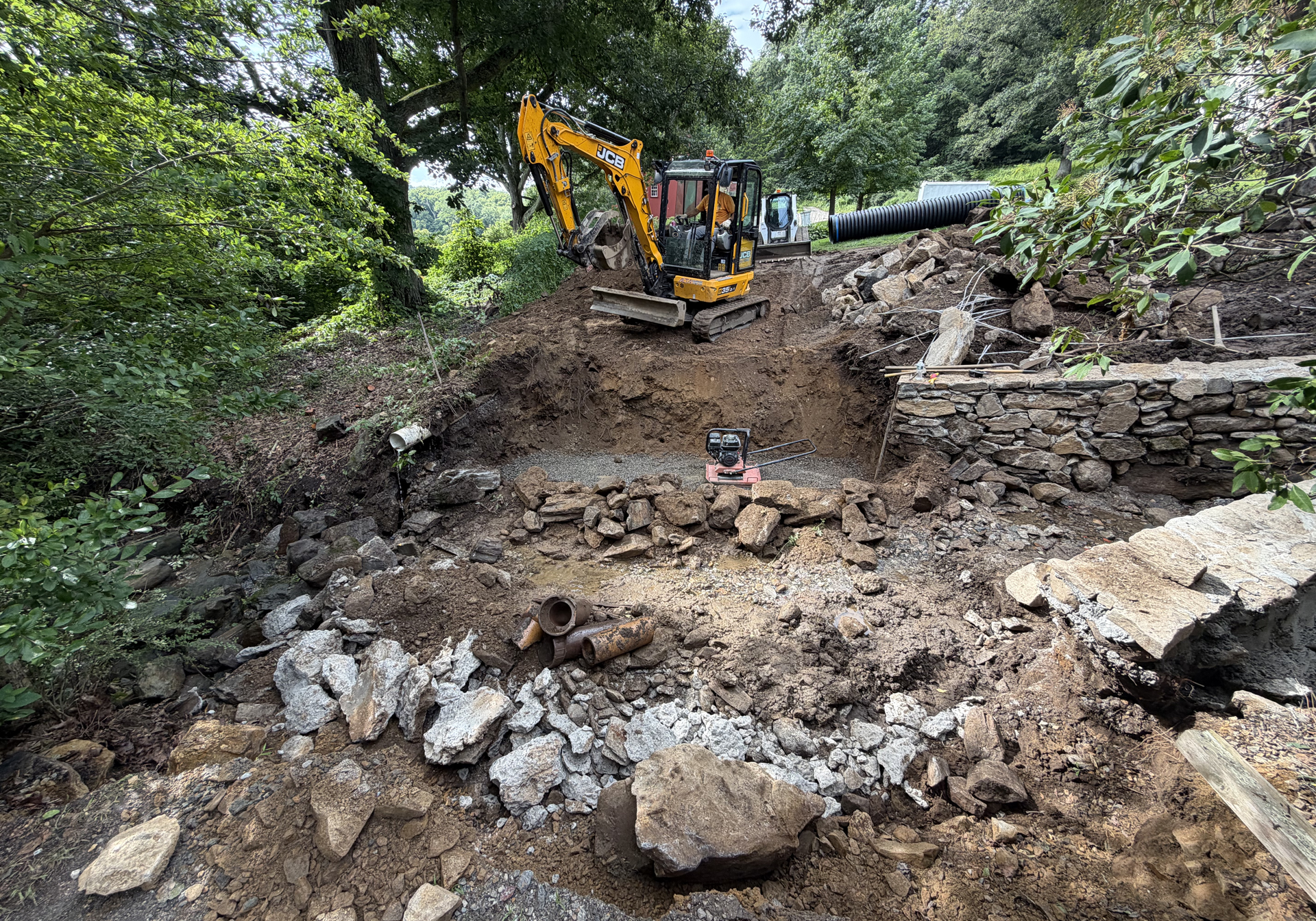 Excavator clearing a muddy, rocky path beside a stone wall in a wooded area