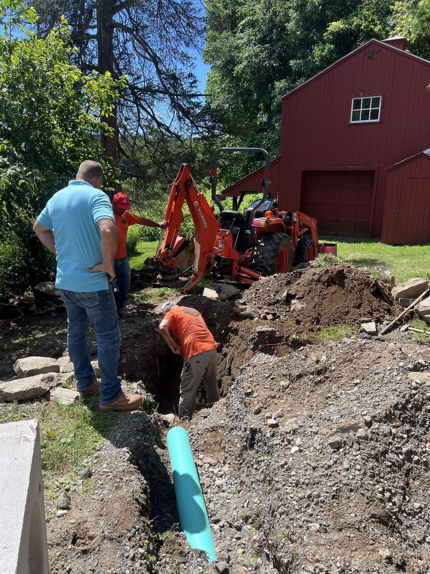 Workers using an excavator beside a red barn, digging a trench in a wooded yard