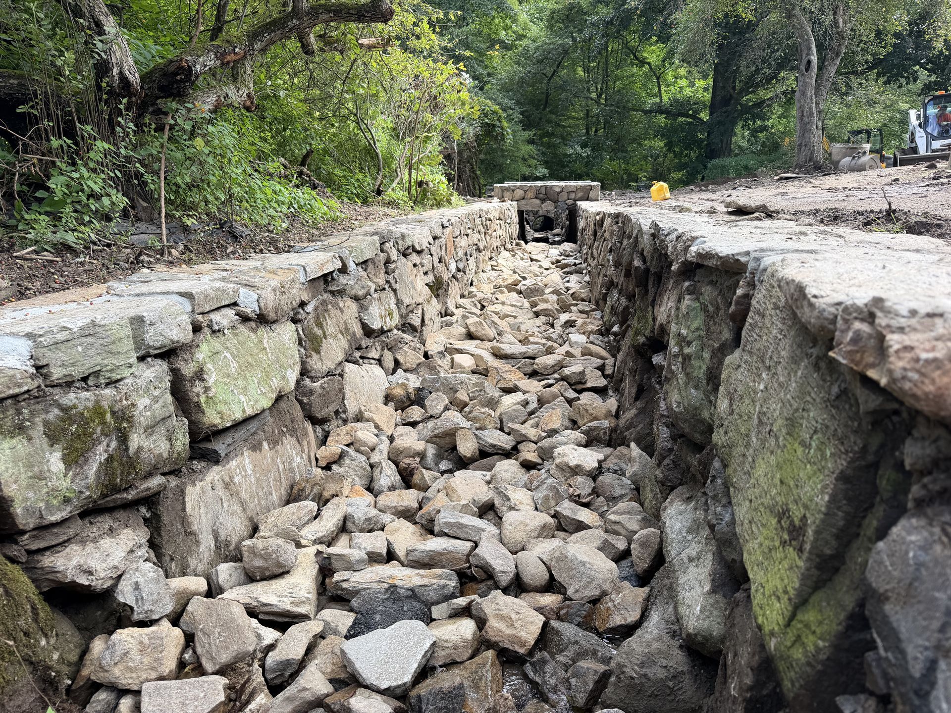 Collapsed stone road in a wooded area, with a deep gully of loose rocks between broken pavement edges