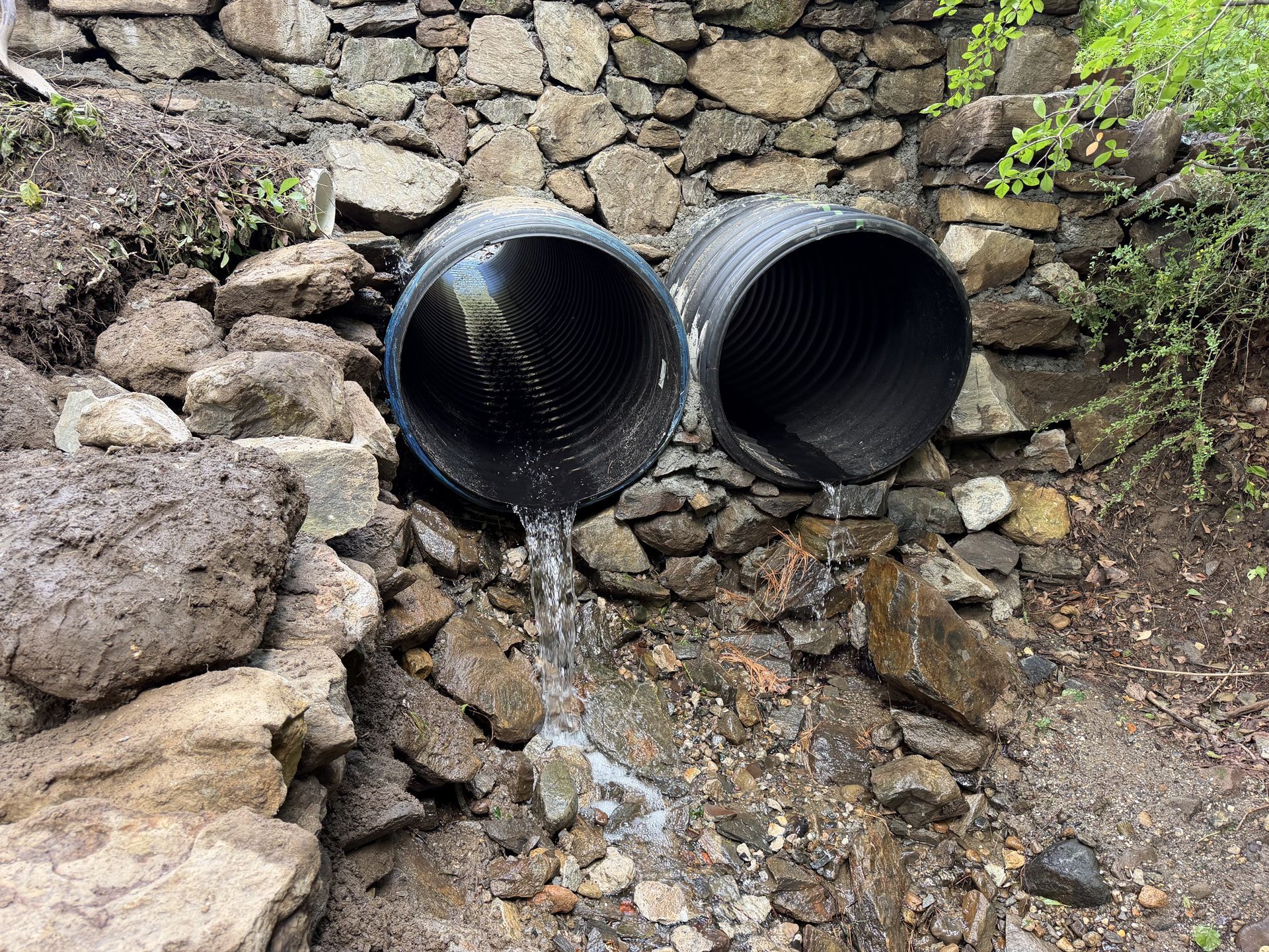 Two black drainage pipes protruding from a stone wall, with water trickling onto rocks below