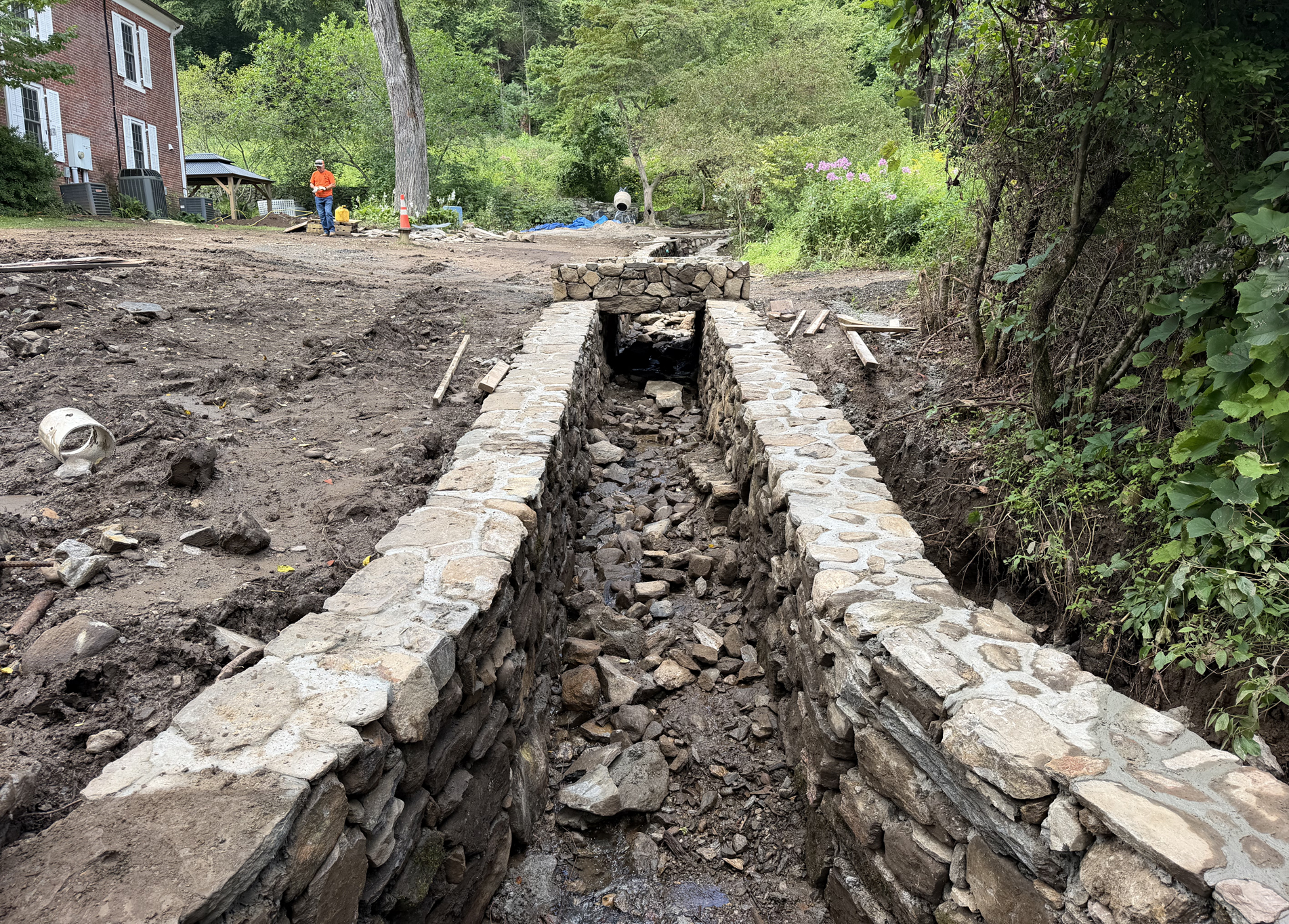 Stone-lined drainage trench under construction beside a wooded path, with excavated soil and a worker in the distance