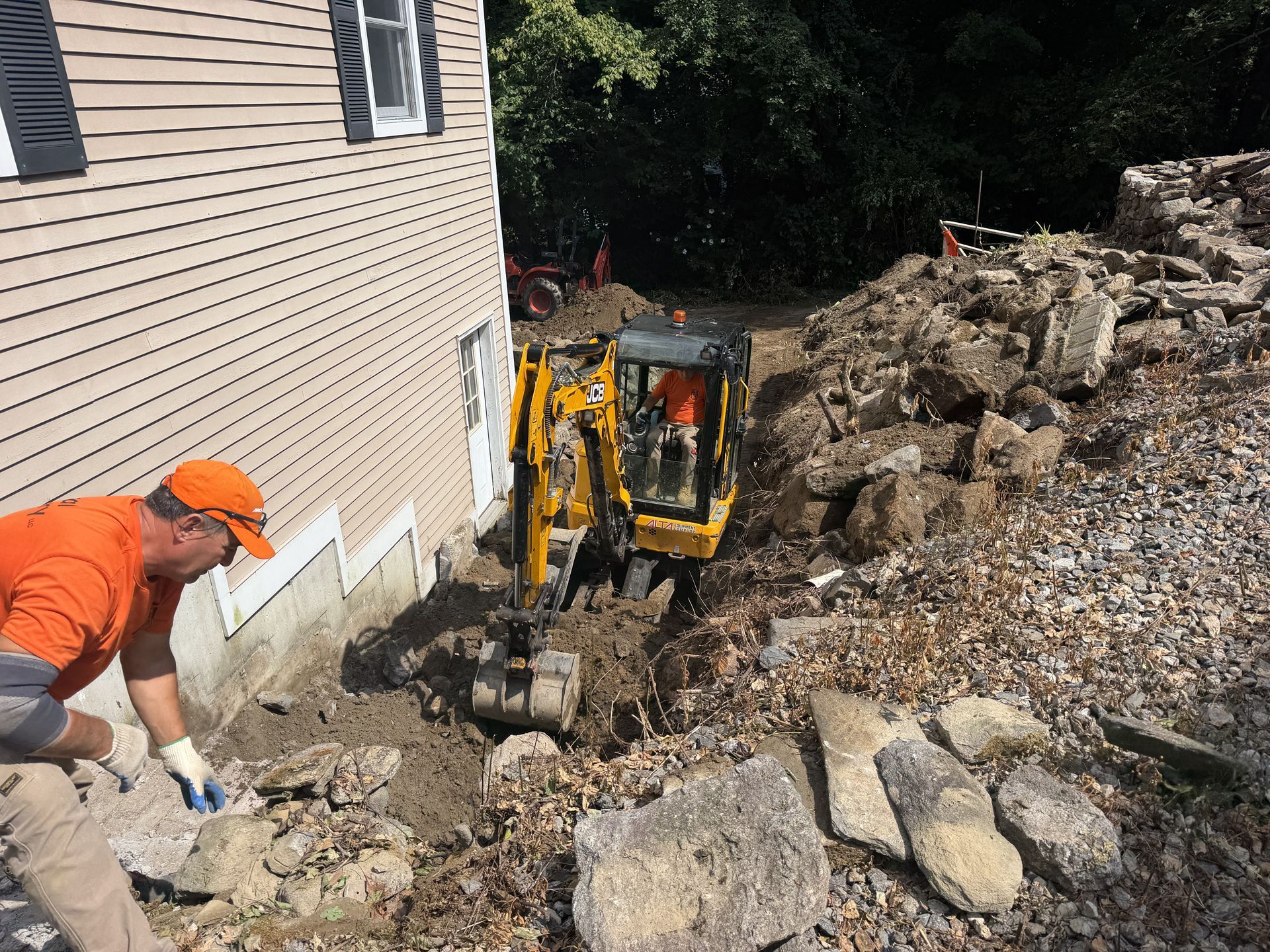 Worker in orange gear operates a small excavator beside a house during excavation work