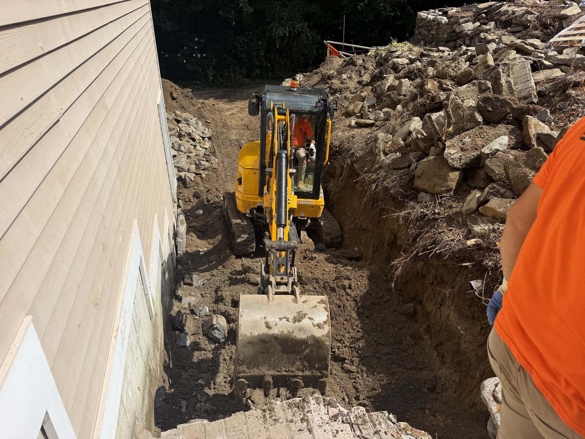 Yellow excavator digging a trench beside a house, with a worker in orange vest at the edge.