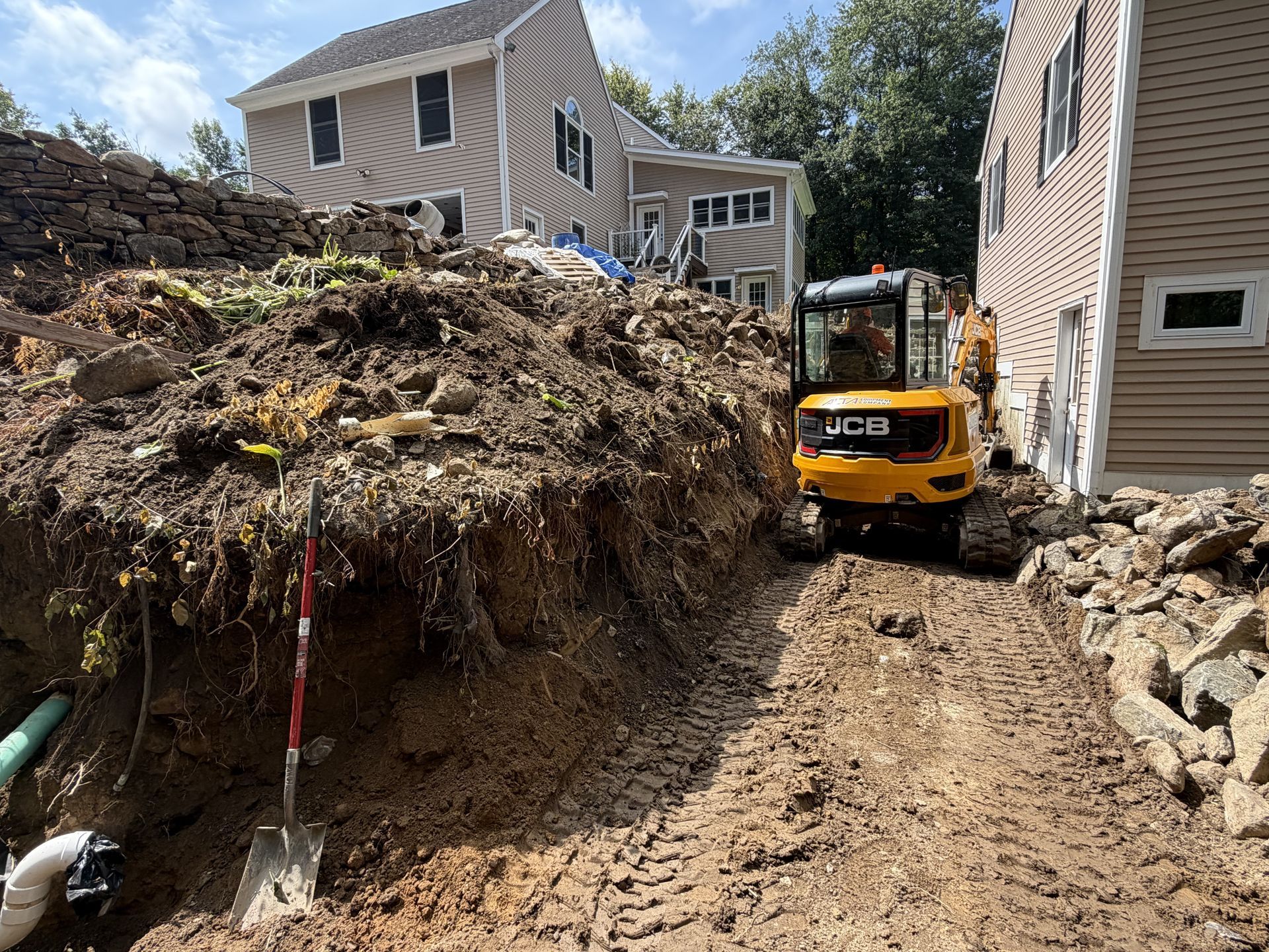 Construction excavator in a narrow trench beside houses, with exposed soil and stone retaining walls.
