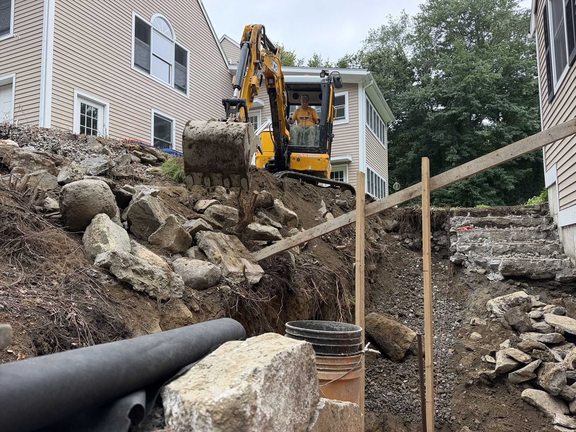 Excavator works on a rocky slope beside two houses under construction, with wooden braces and pipes in foreground.