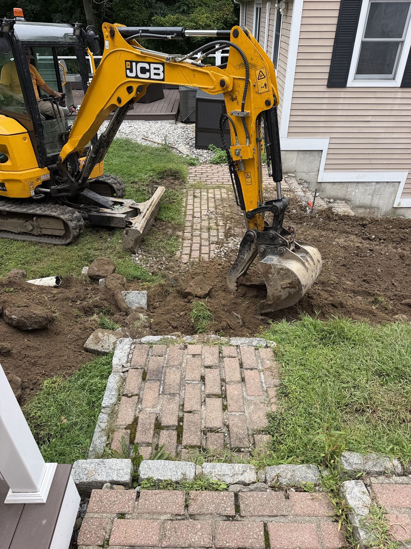 Yellow JCB mini excavator digging a yard beside a brick walkway and house siding