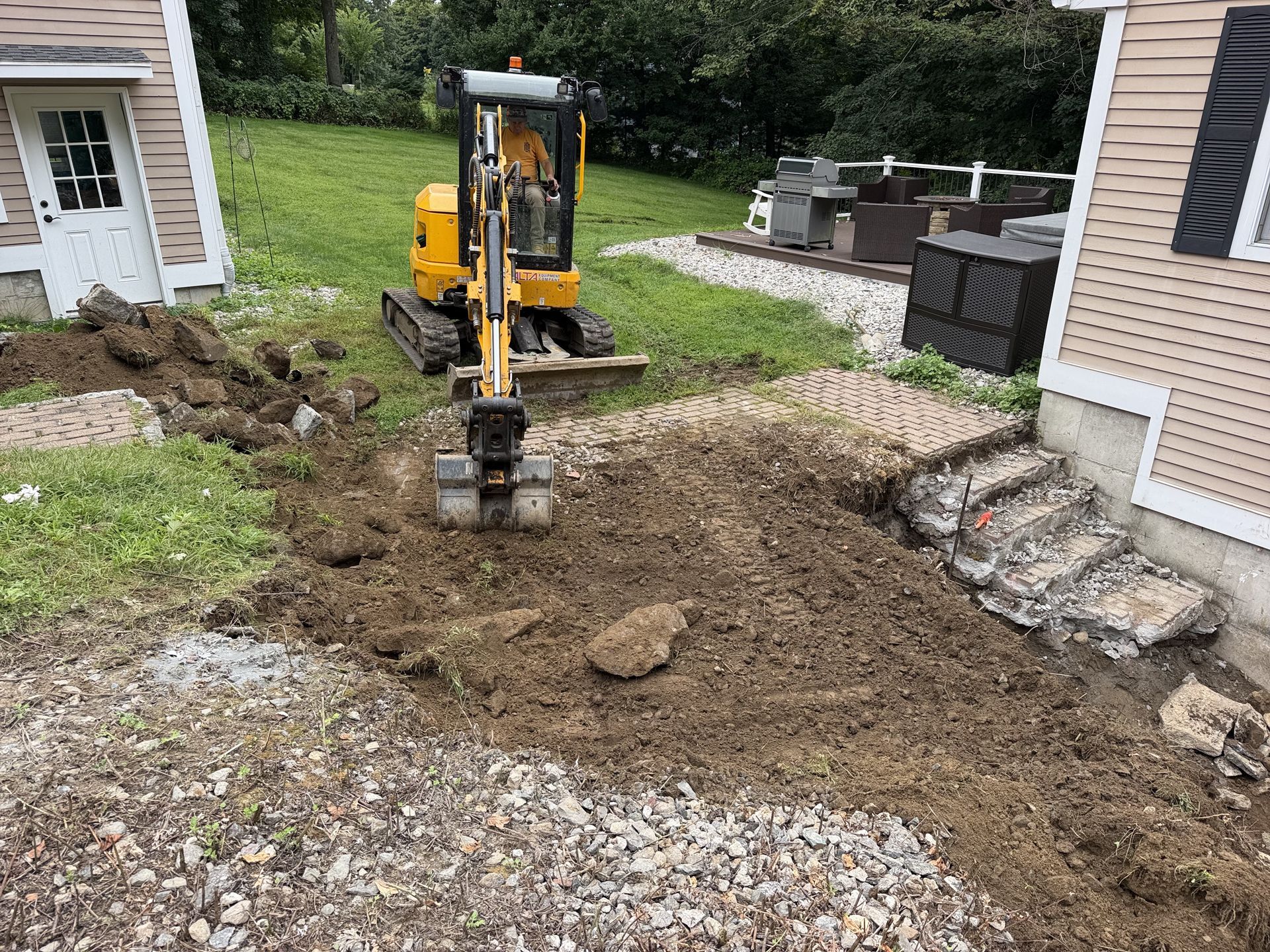 Mini excavator digging dirt beside a house and stone patio in a backyard.