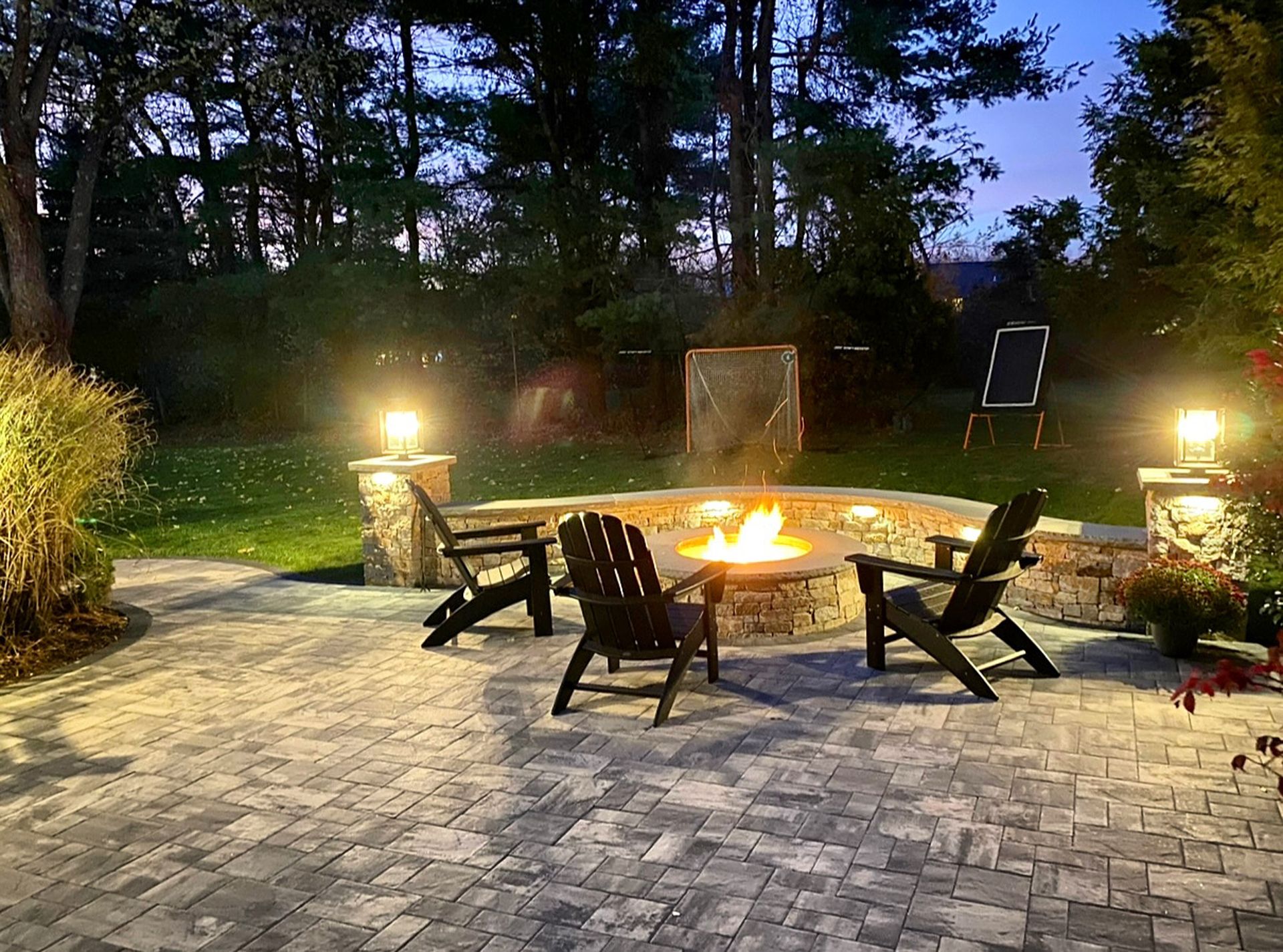 Backyard patio with stone fire pit, Adirondack chairs, and warm lights at dusk