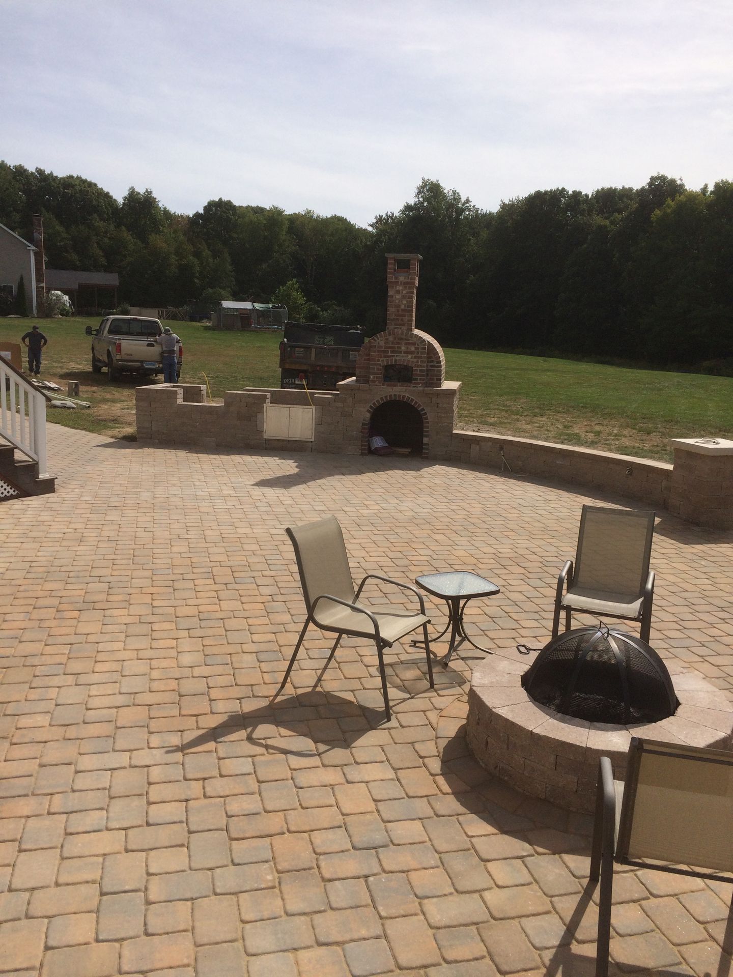 Patio with stone grill, fire pit, and chairs overlooking a grassy yard and trees
