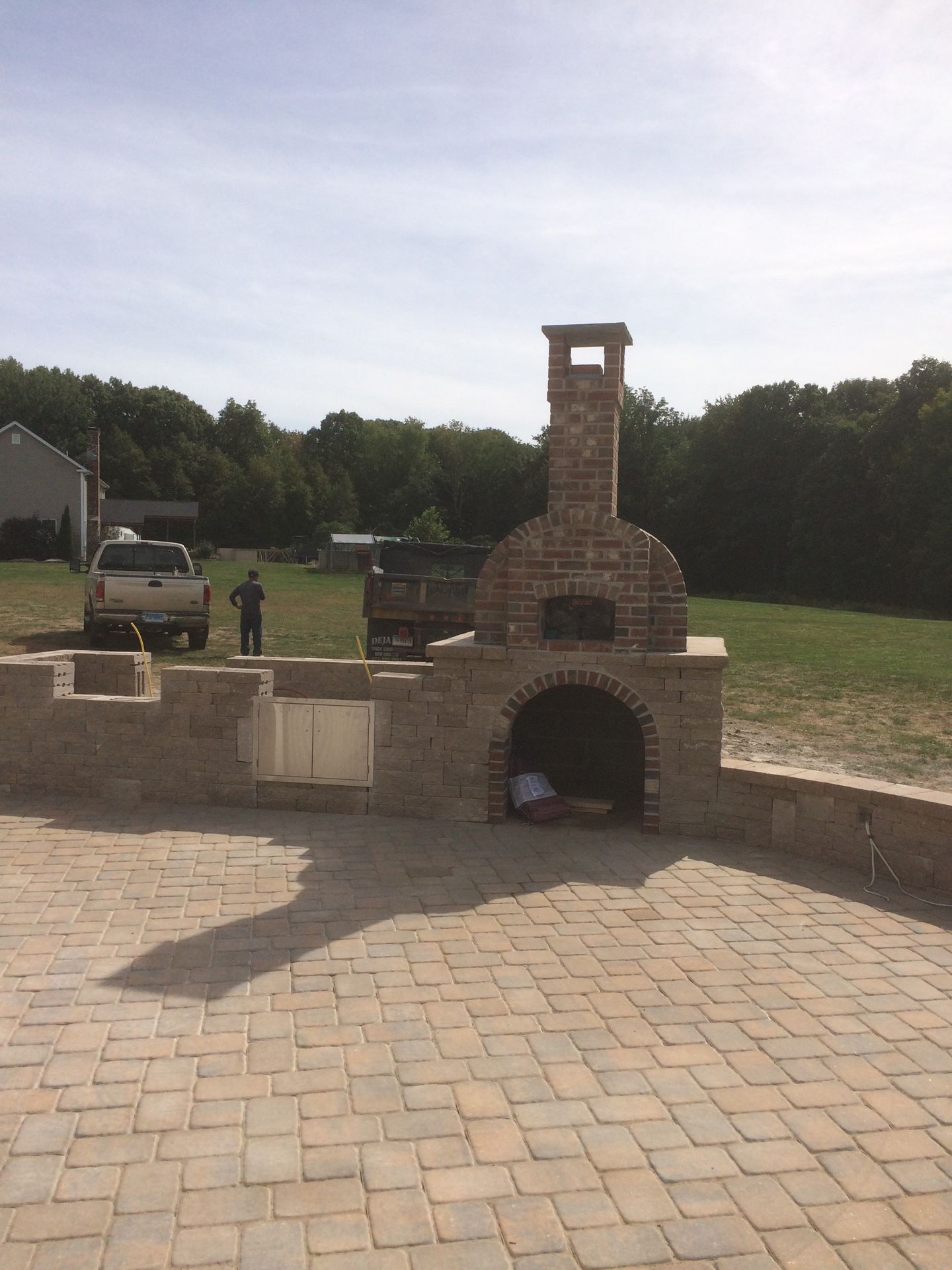 Brick outdoor oven and chimney on a paved patio beside a grassy field, with vehicles in the background