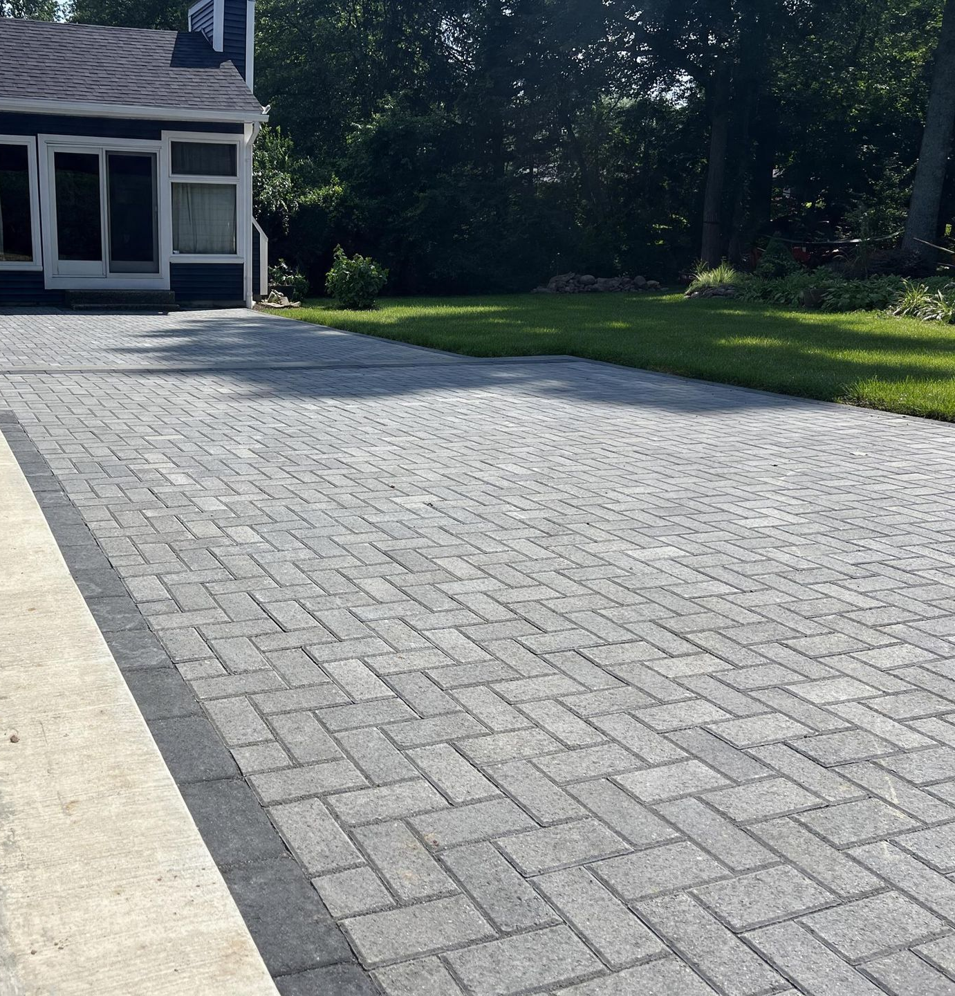 Stone paver patio beside a house, with green lawn and trees in the background.