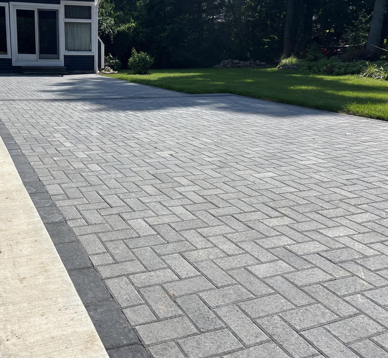 Gray brick-paved driveway beside a concrete path and green lawn in front of a house