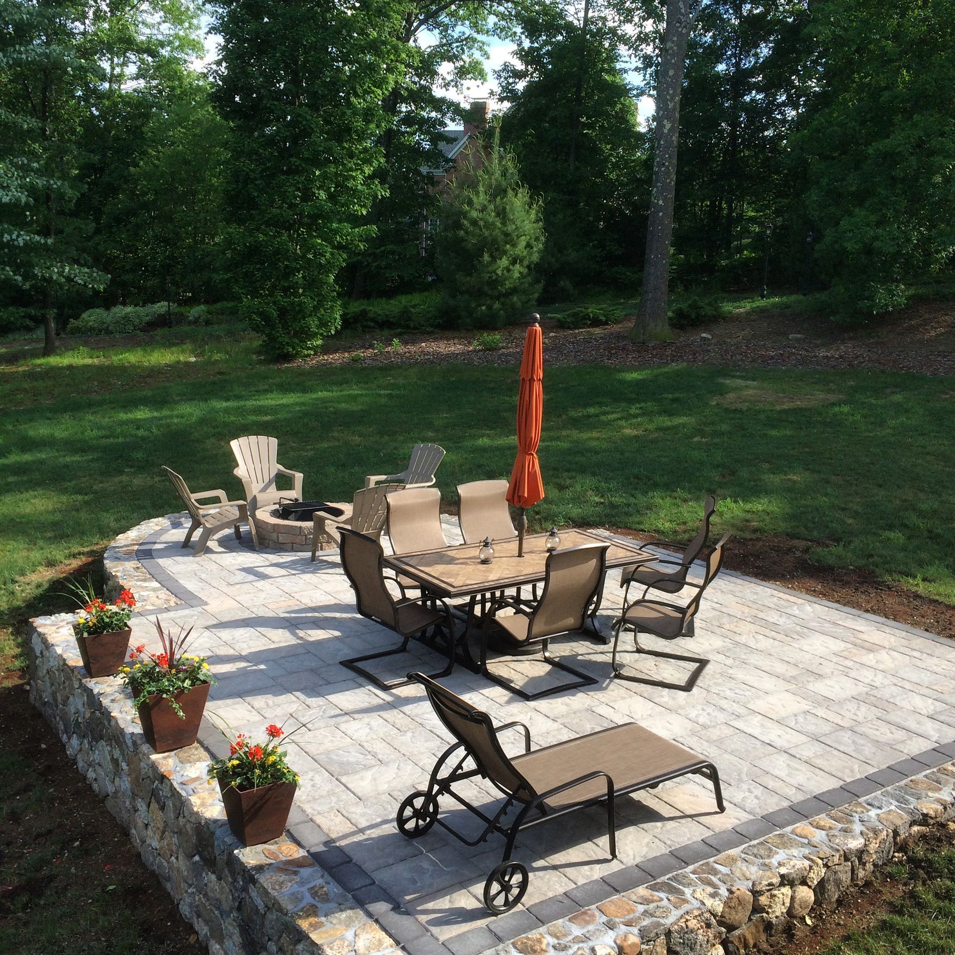 Backyard patio with outdoor dining table, chairs, lounge chairs, and a closed orange umbrella on stone pavers