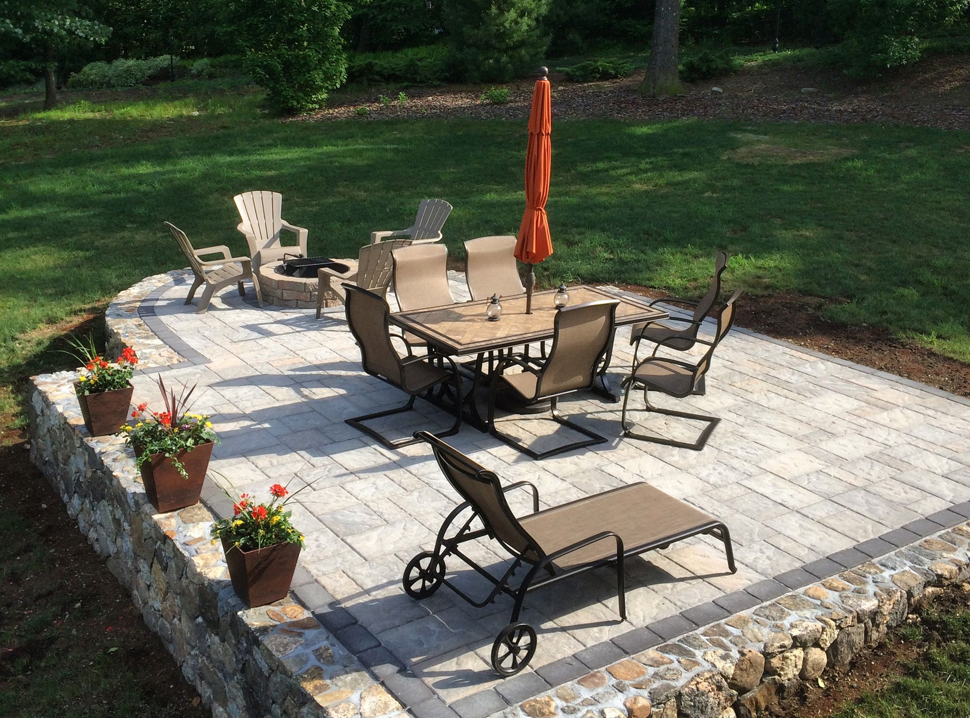 Patio with outdoor seating, tan lounge chairs, potted flowers, and a closed orange umbrella on a stone patio.