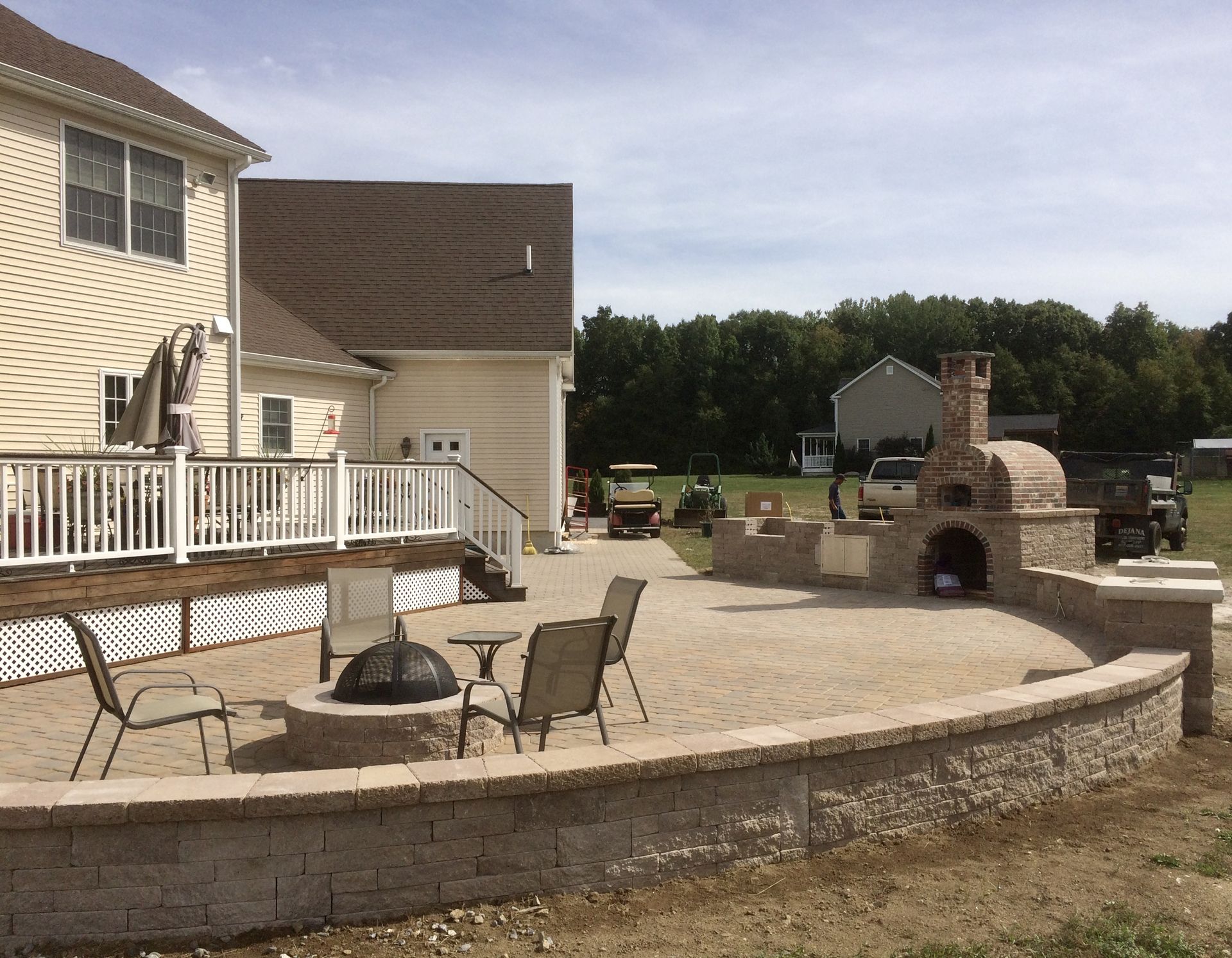 Patio with outdoor seating and stone fireplace beside a house, overlooking a grassy yard and trees
