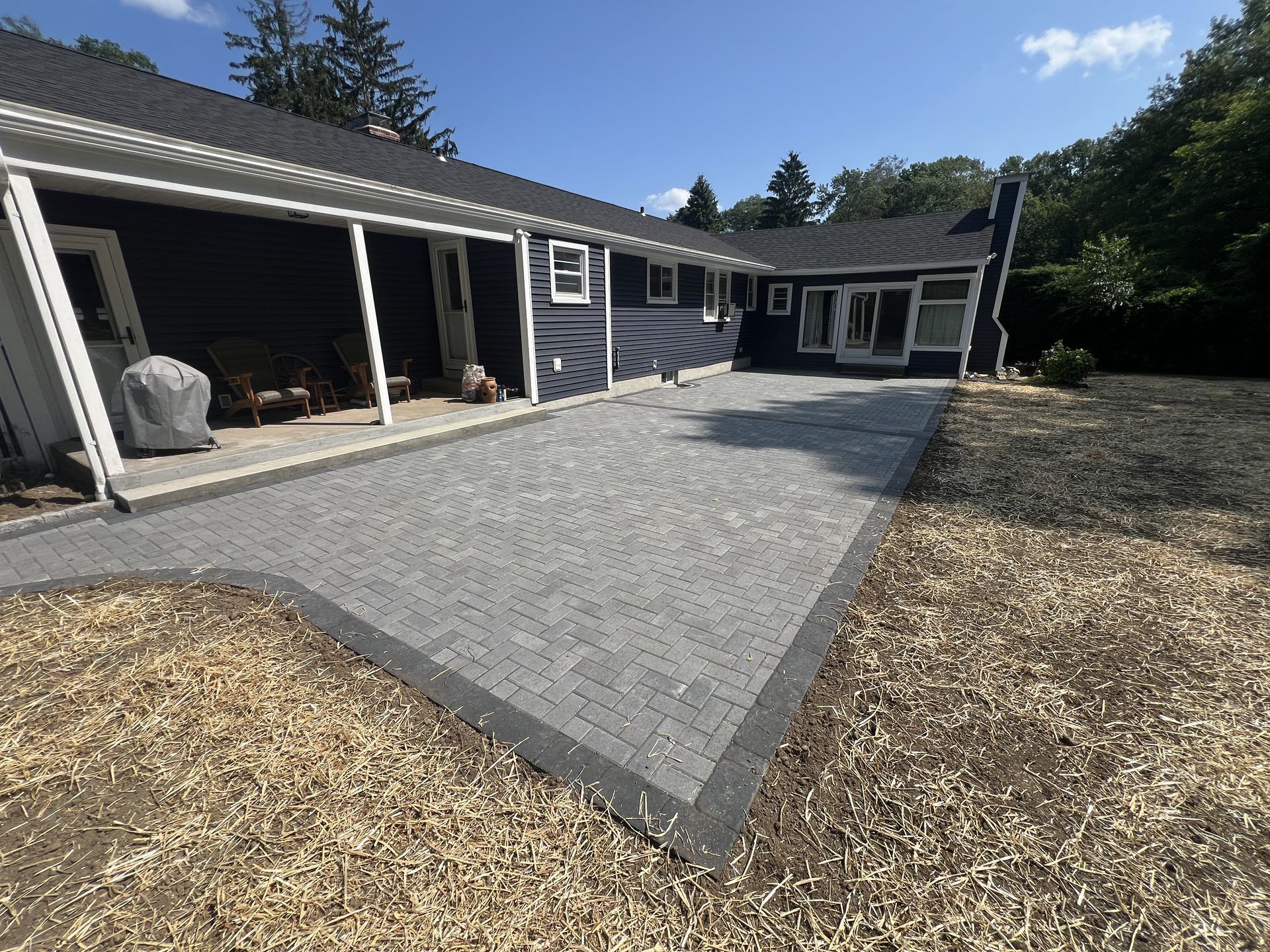 Gray paver patio beside a house with covered seating and wood-chip landscaping under a blue sky