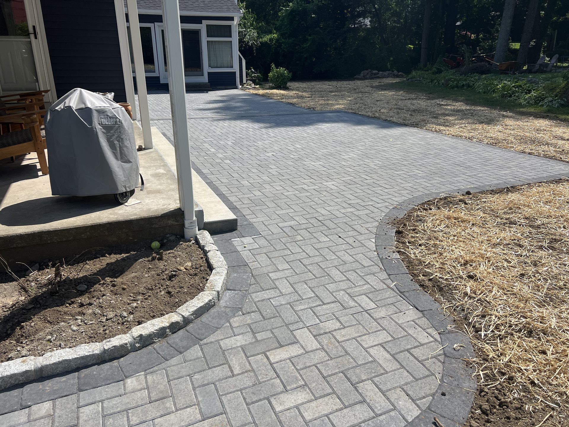 Curved gray paver walkway leading to a gravel driveway beside a house and covered grill